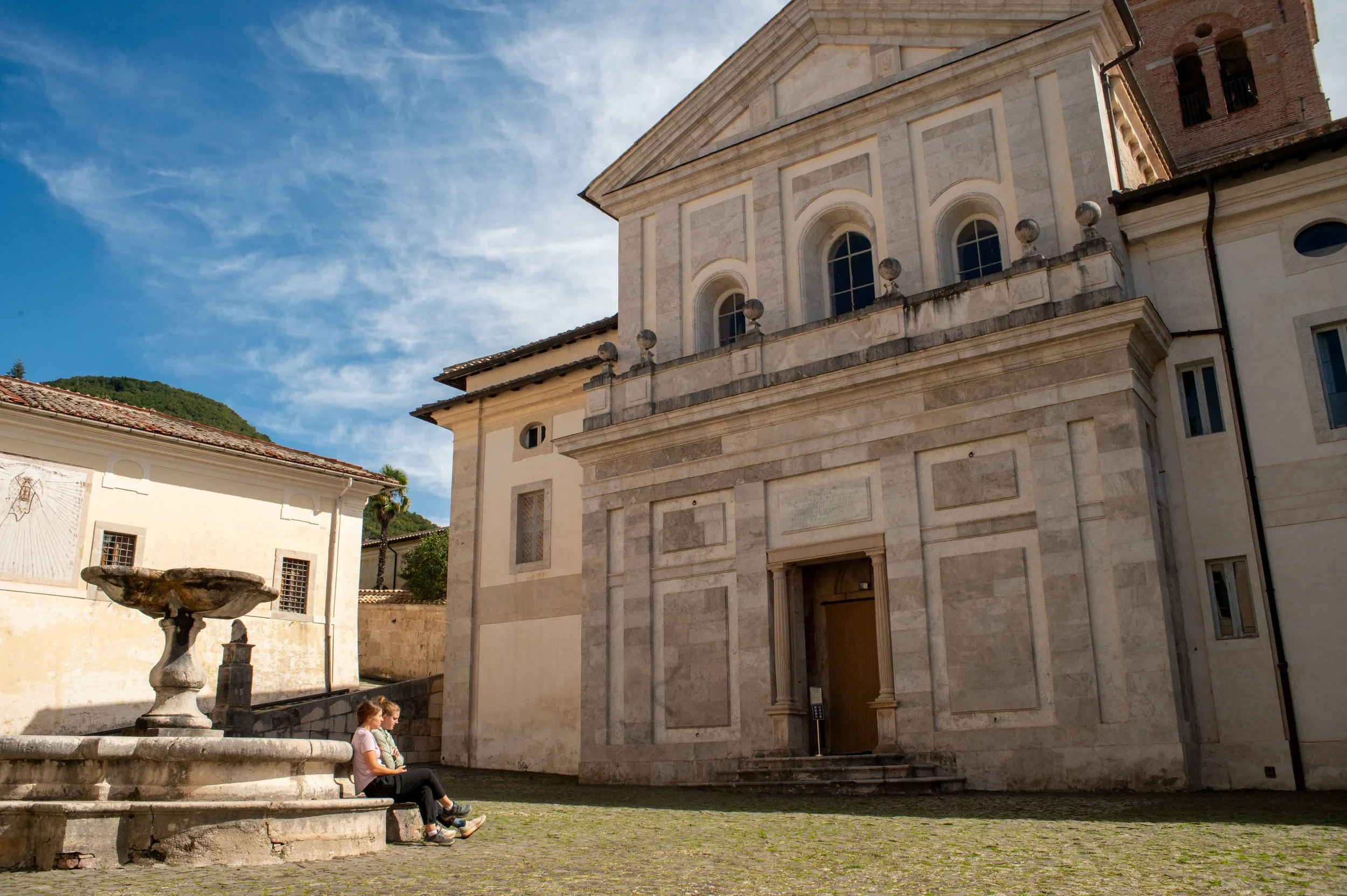 Two people sitting on a stone bench near a historic European church with a fountain and cobblestone walkway under a clear blue sky.