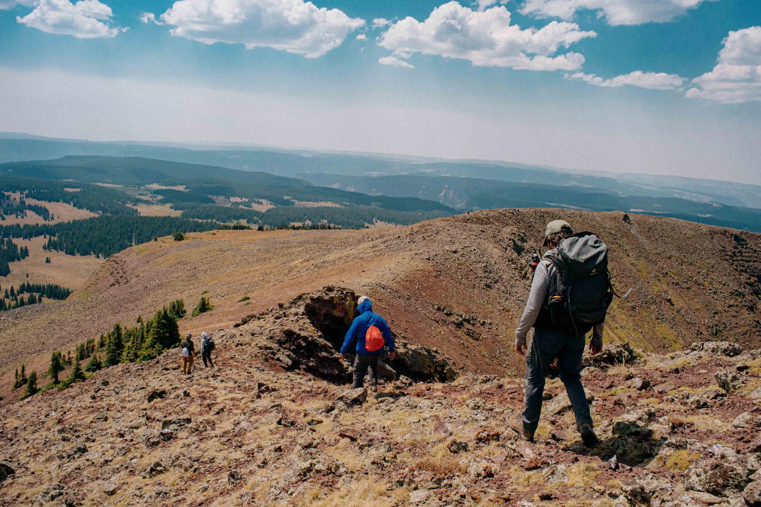 Four hikers with backpacks walking on a rocky mountain trail under a partly cloudy sky, with forested and open rolling hills in the distance.