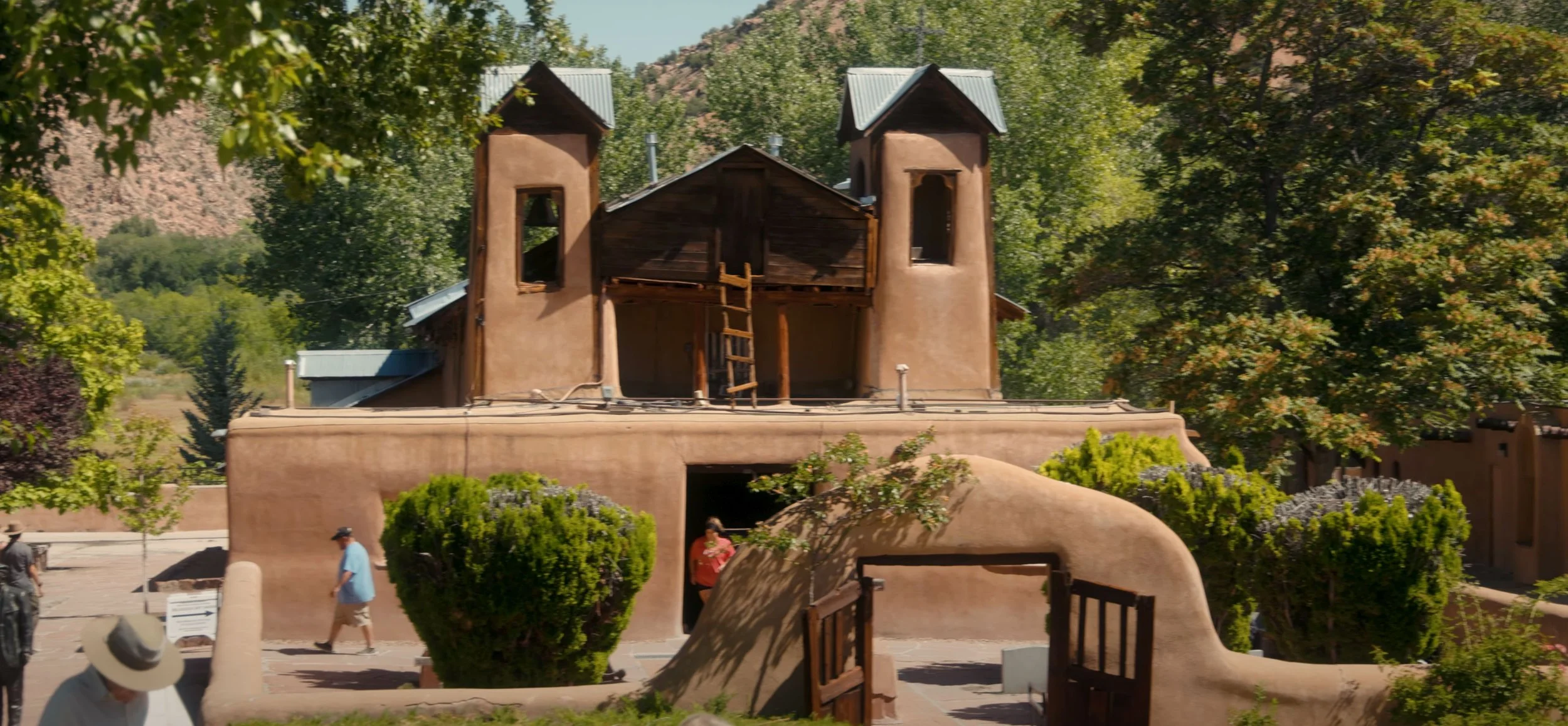 A traditional adobe-style building with two towers and a courtyard, surrounded by trees and mountains in the background, with people walking nearby.