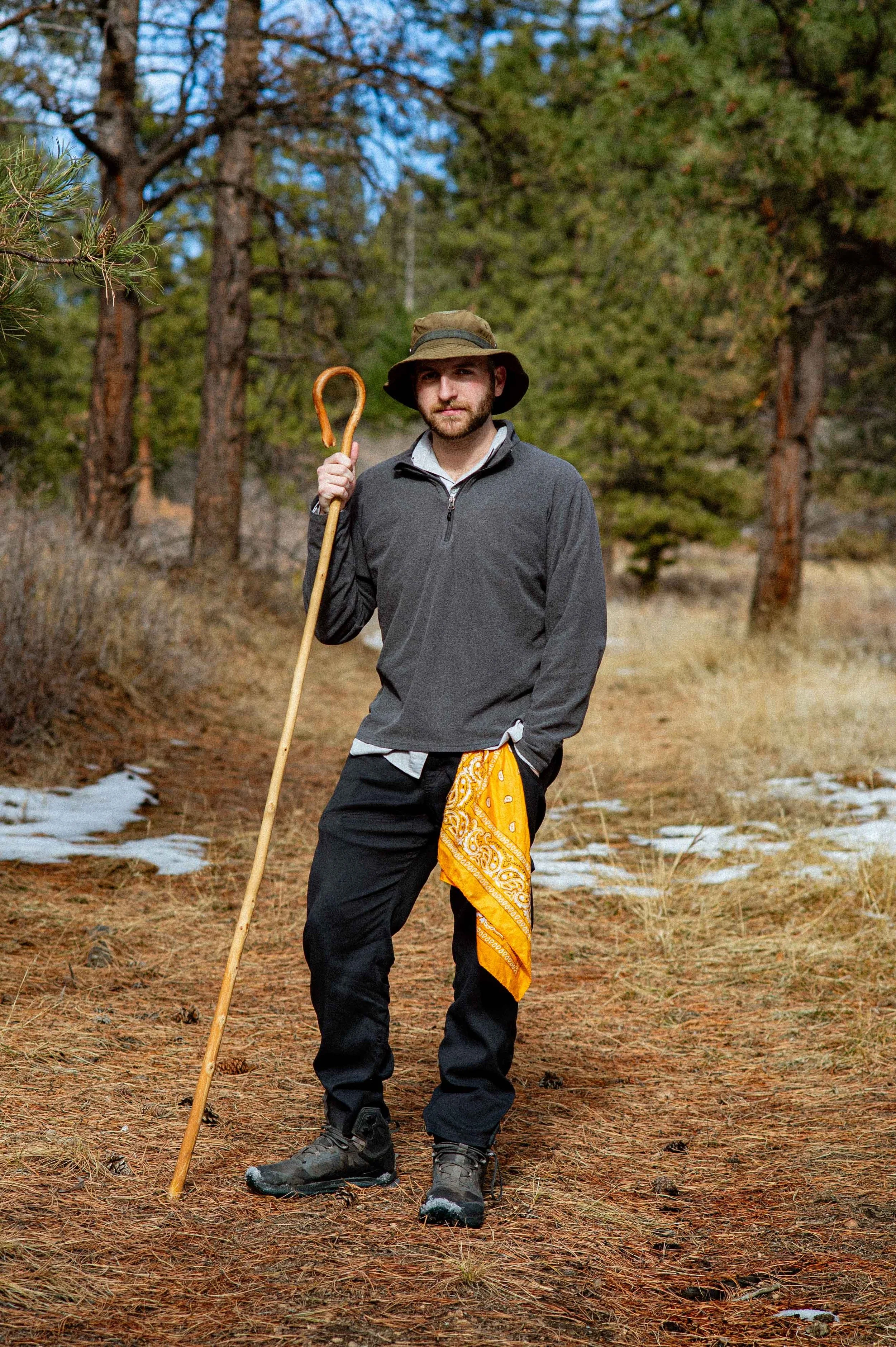 A man standing outdoors in a wooded area holding a walking stick, wearing a hat, gray sweater, black pants, and hiking boots.