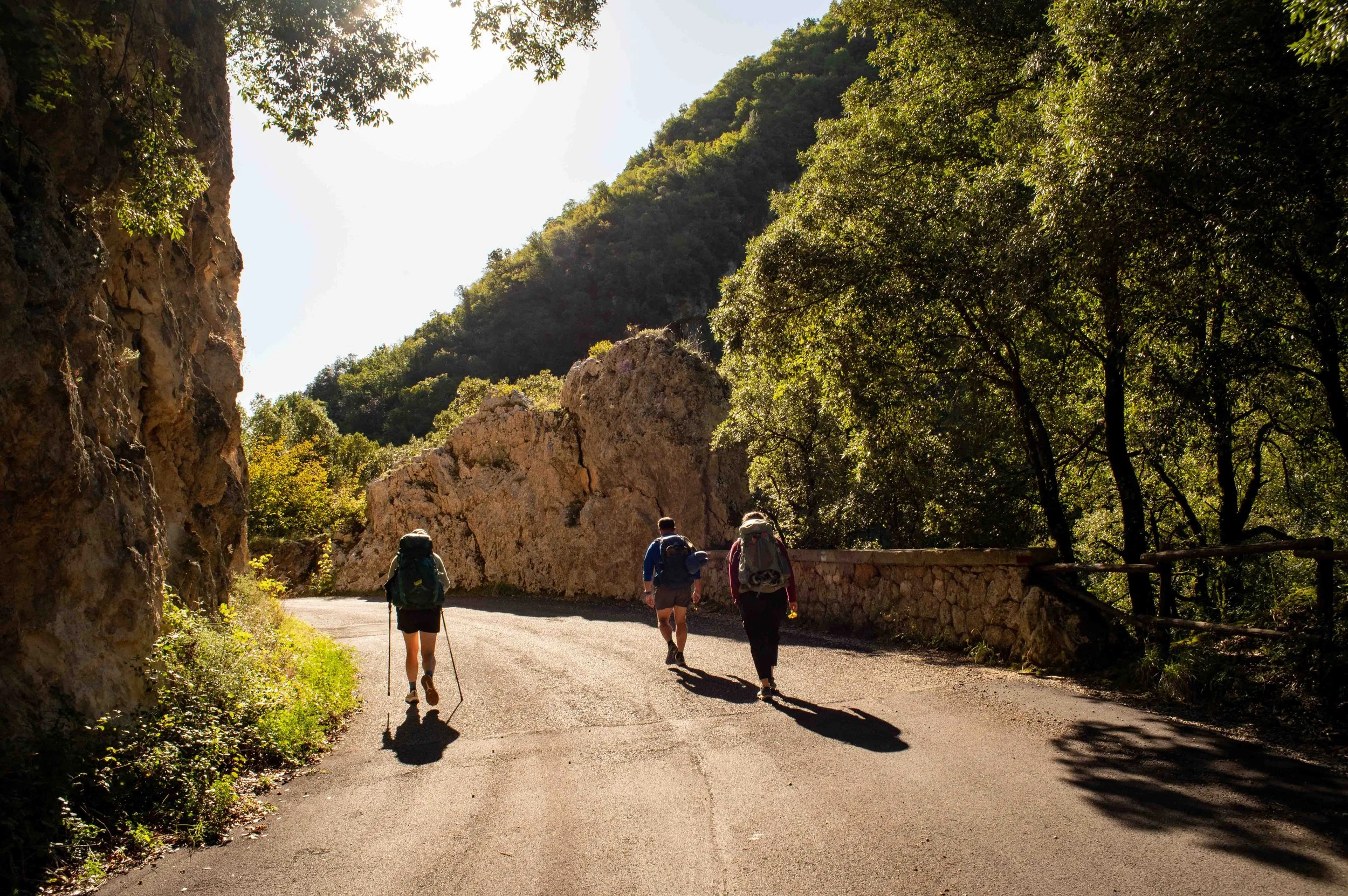 Three hikers walking along a mountain road surrounded by trees and rocky cliffs under sunlight.