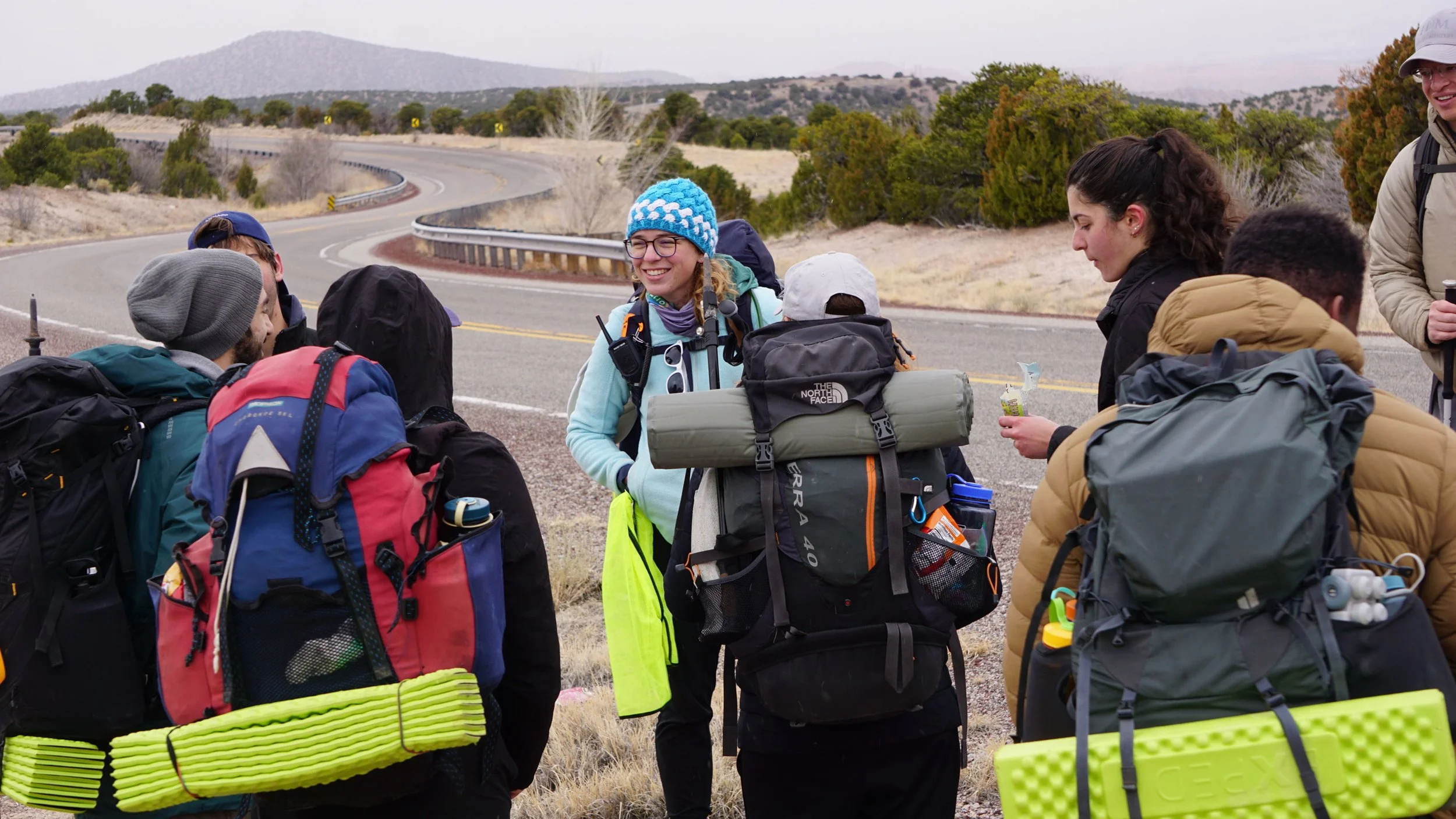 Group of hikers with backpacks gathered on side of a winding road in a rural, mountainous area with sparse vegetation and cloudy sky.