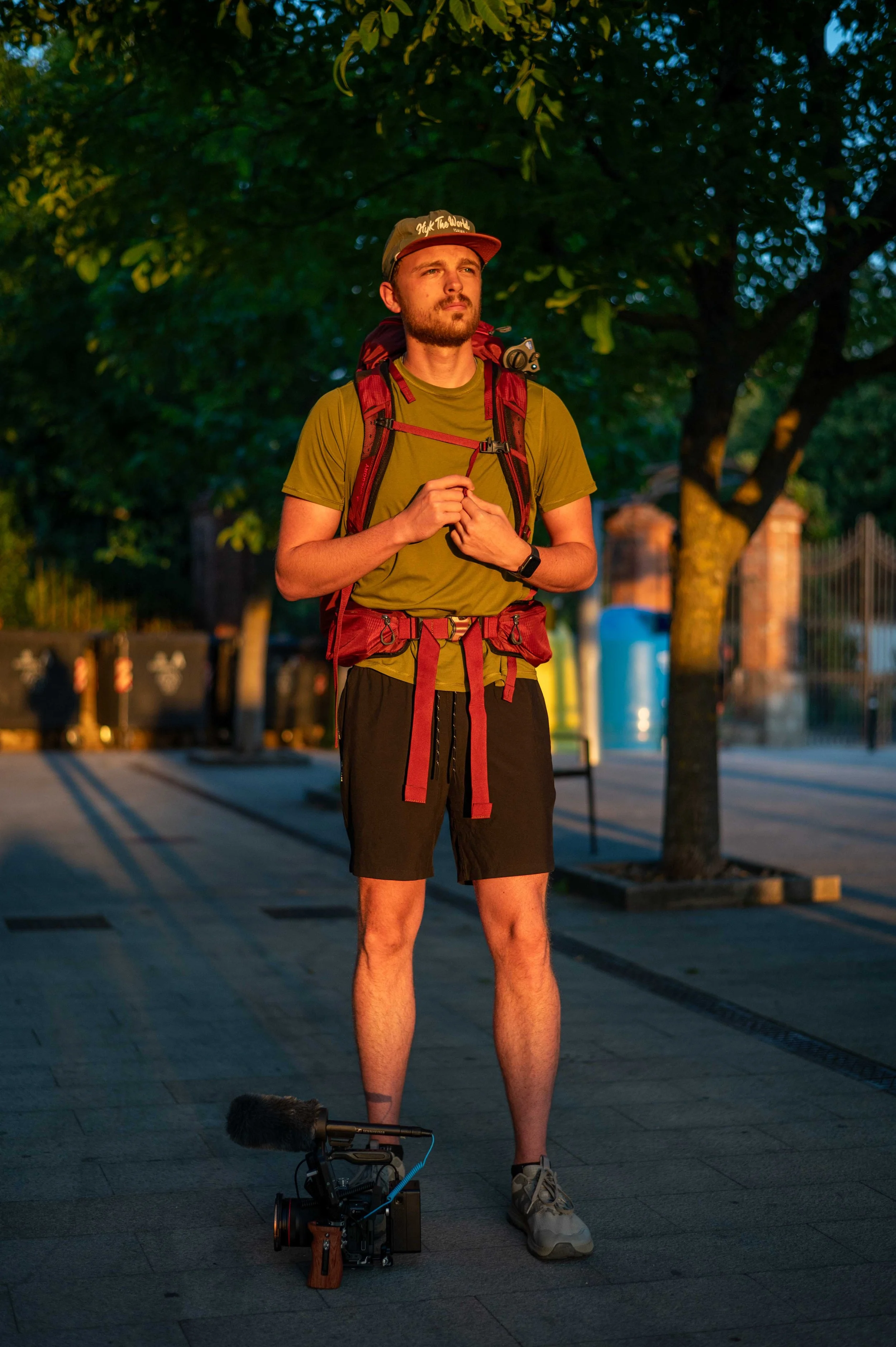 A man with a beard wearing a green t-shirt, black shorts, sneakers, and a cap, standing outdoors at dusk with a backpack and a camera with a microphone on the ground beside him.