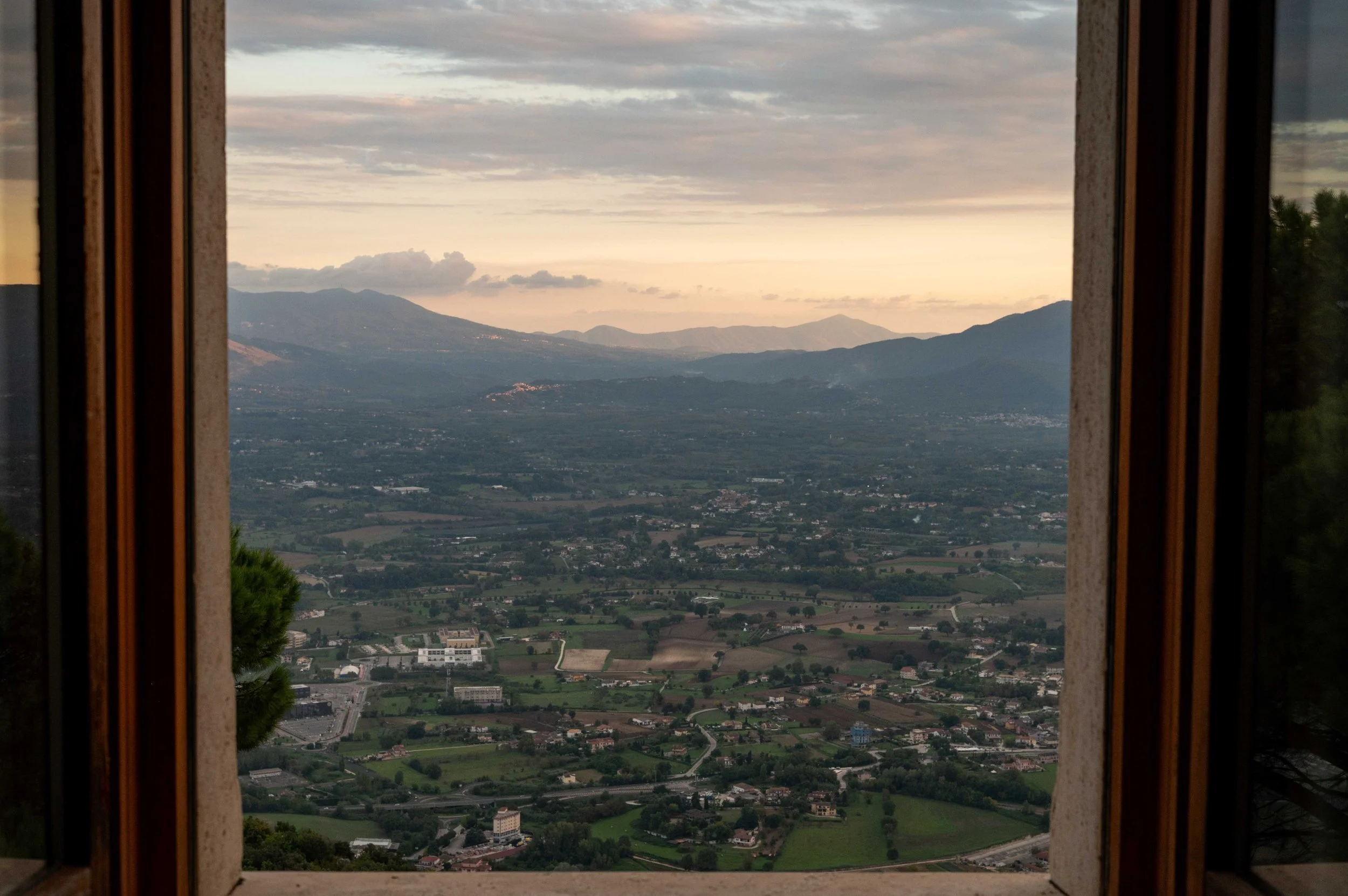 View of a valley with fields, buildings, and roads, framed by open window with mountains in the distance and a cloudy sky.