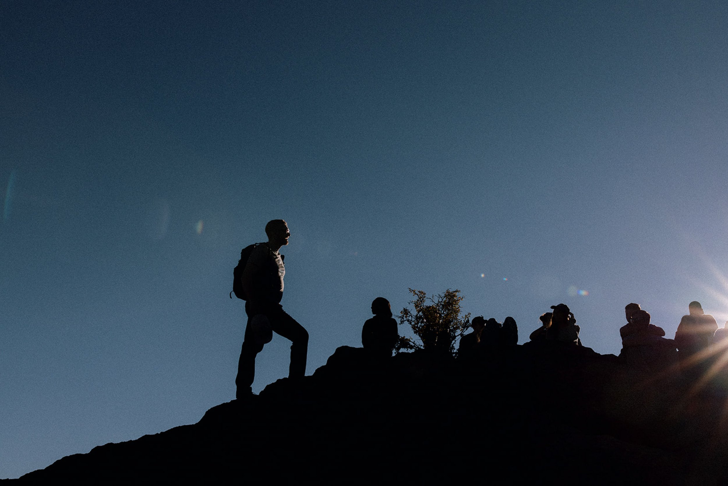 People sitting on a rocky hilltop with one person standing and others sitting in the sunset or sunrise, with a clear sky and sun rays on the horizon.