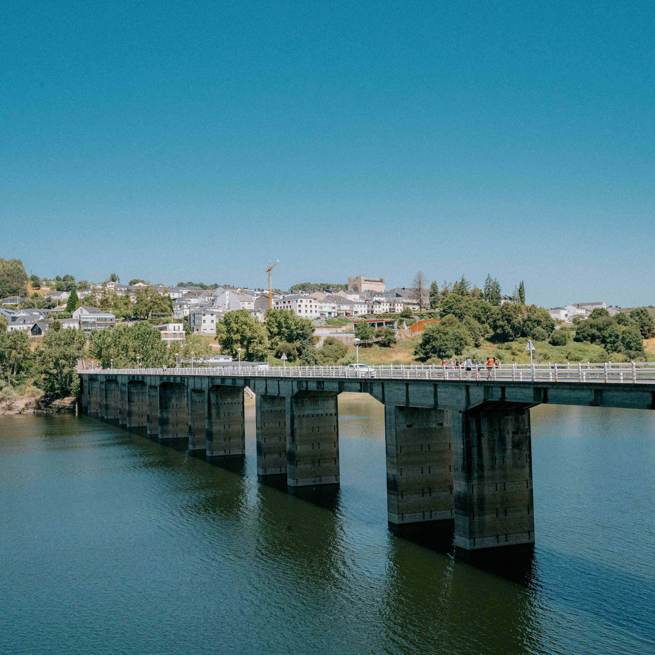 A bridge crossing a river with a town on a hill in the background under a clear blue sky.
