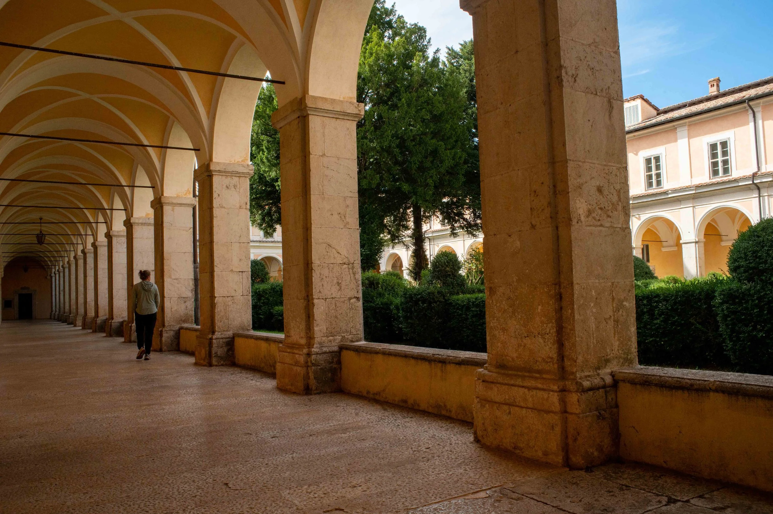 A woman walking through a covered walkway with arches on the left side and a view of a courtyard with green bushes and trees on the right side, under a blue sky.