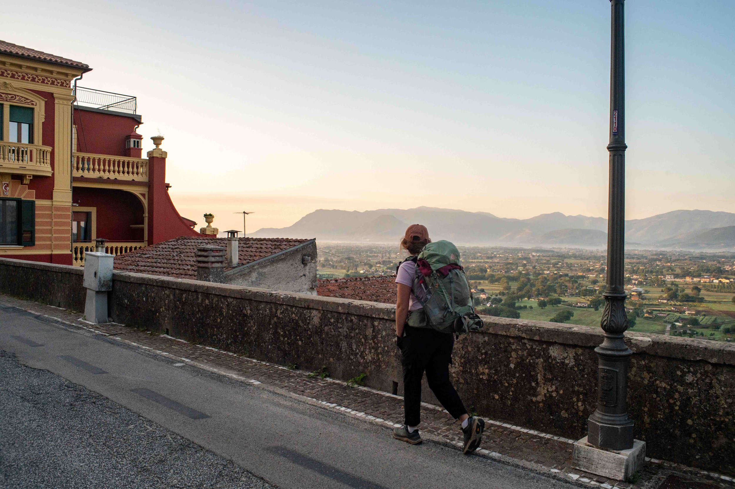 Hiker with a backpack walking along a city street at sunset, overlooking a valley with mountains in the distance.