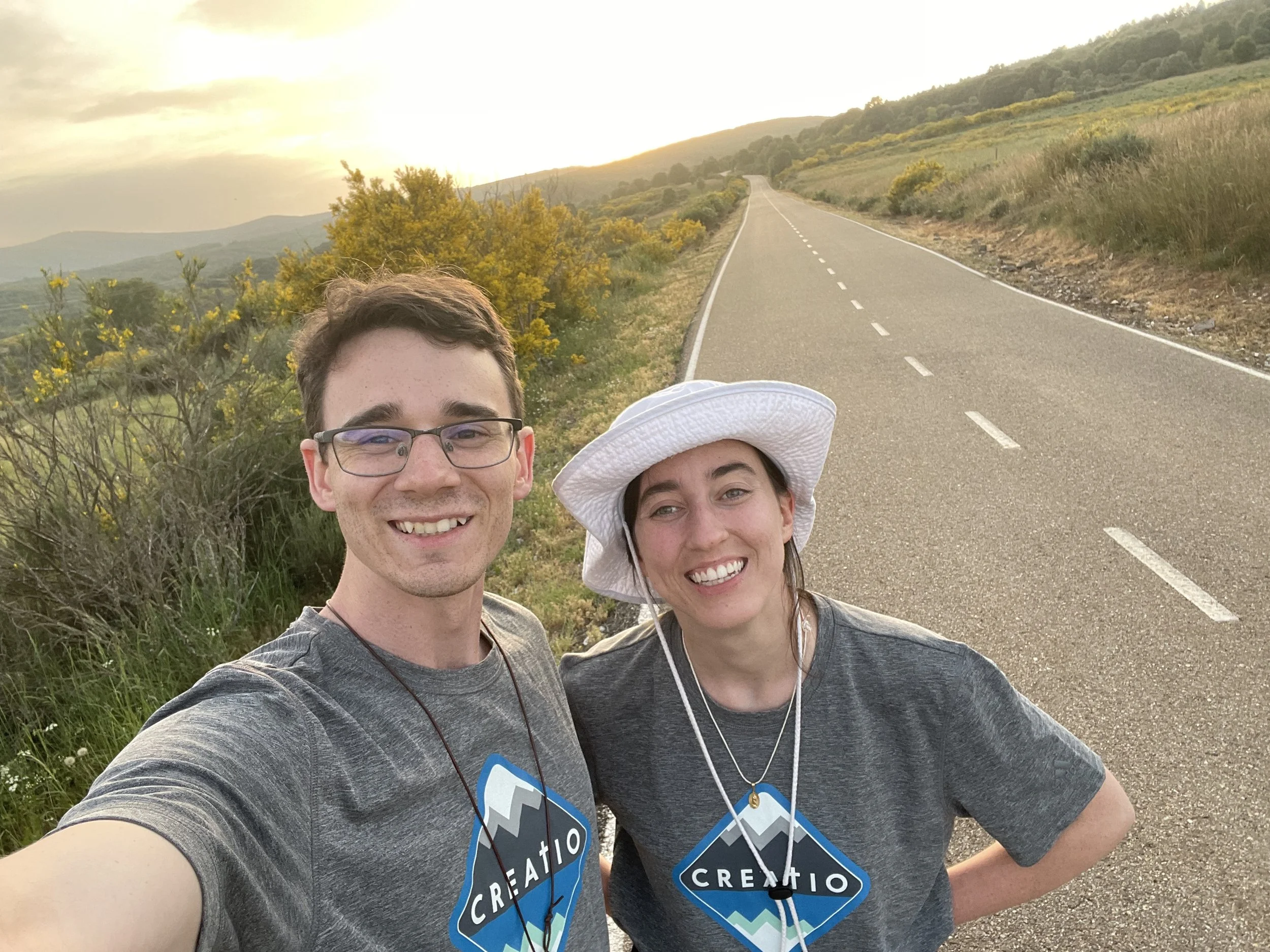 A smiling young man and woman taking a selfie on a rural road at sunset, both wearing gray T-shirts with a mountain-themed logo and white sun hats.