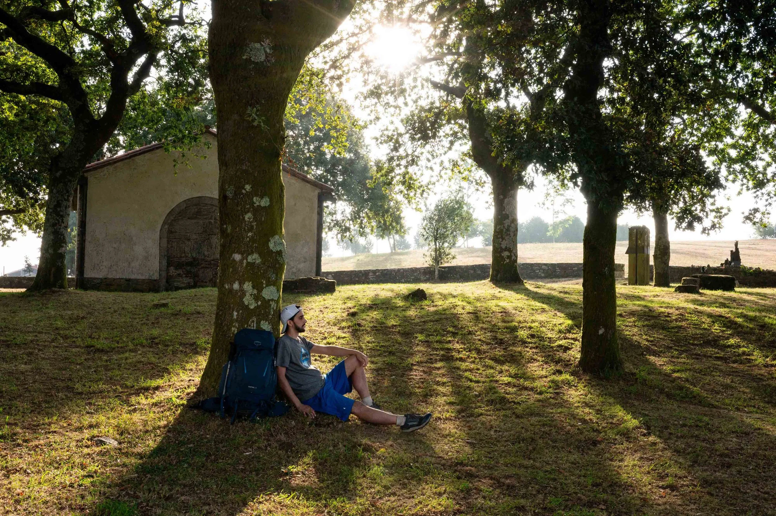 A young man sitting under a large tree during late afternoon or early evening, with a backpack beside him. The scene is in a park or rural area with a small building and a stone monument in the background, sunlight filtering through the leaves.