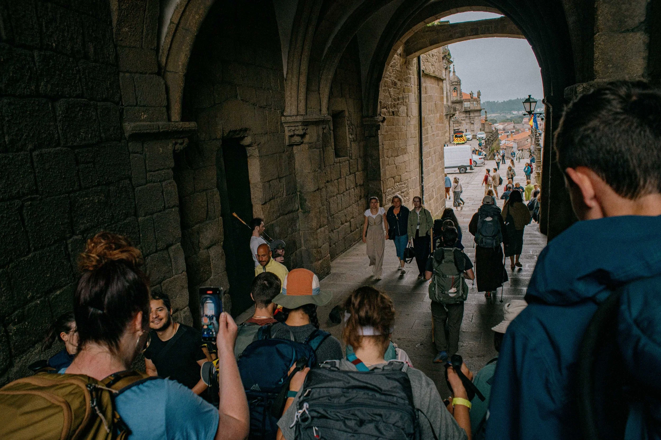 Tourists and visitors walking through an arched stone passageway in a historic city, with some taking photos and others chatting, with the city bustling in the background.