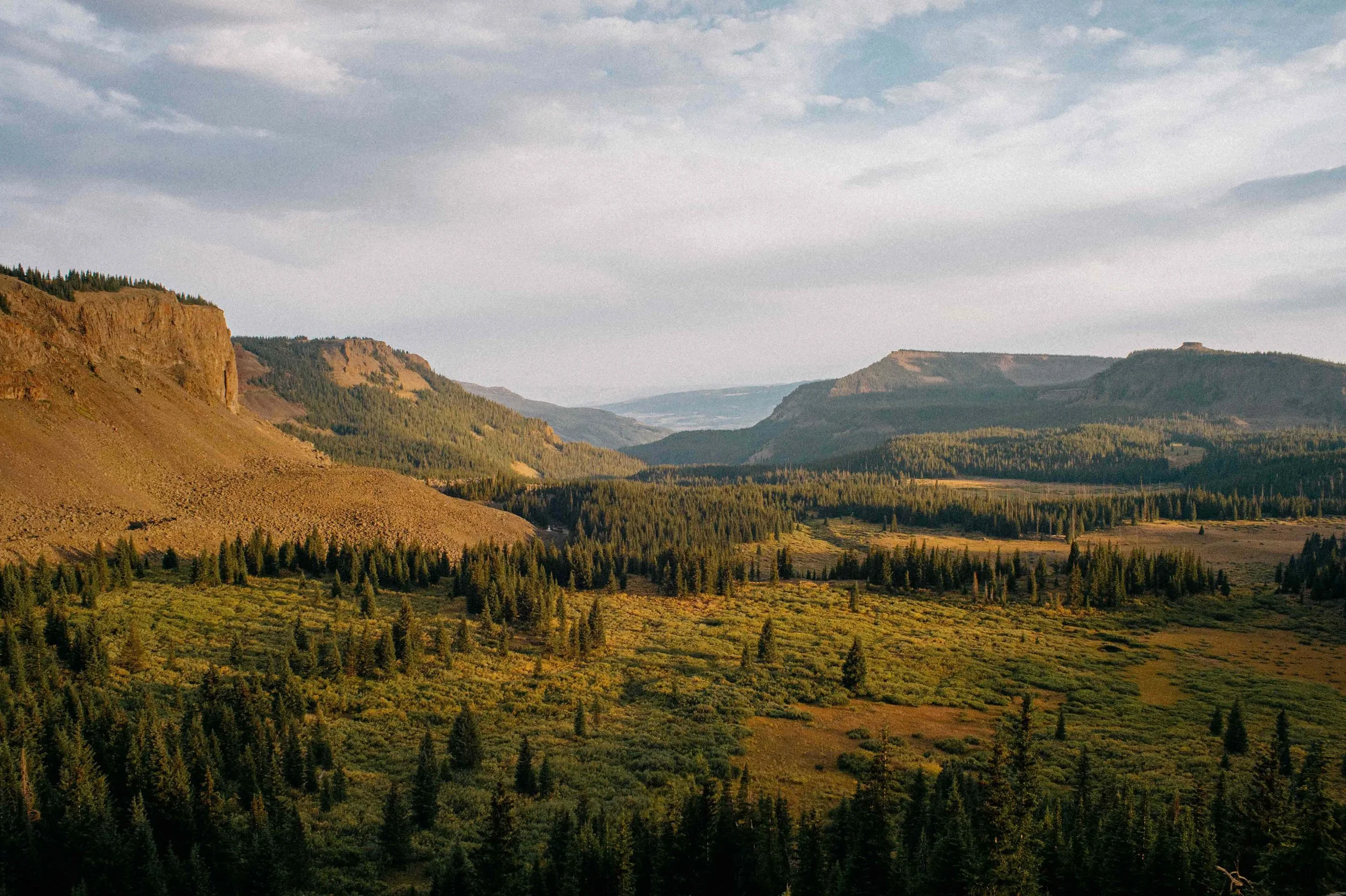 A vast mountain landscape with rolling hills, dense forests, and rocky cliffs under a cloudy sky, bathed in warm sunlight.