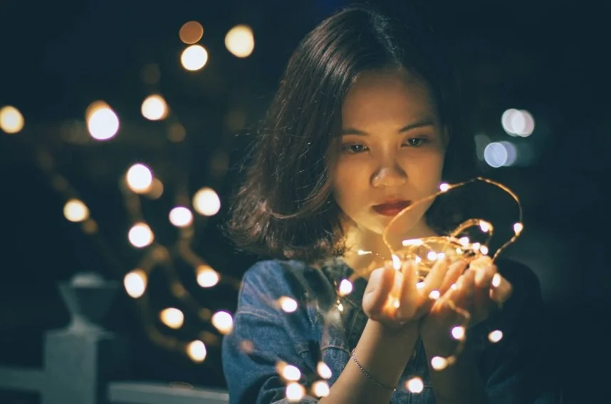 A woman with shoulder-length black hair holding string lights in her hands, looking at them with a thoughtful expression, with blurred lights in the background.