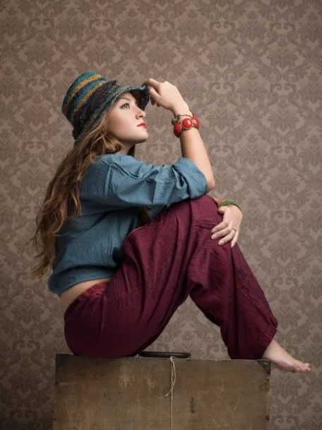 Young woman sitting on a wooden box, wearing a colorful knit hat, a teal blouse, and maroon pants, with jewelry, against a textured patterned wall.