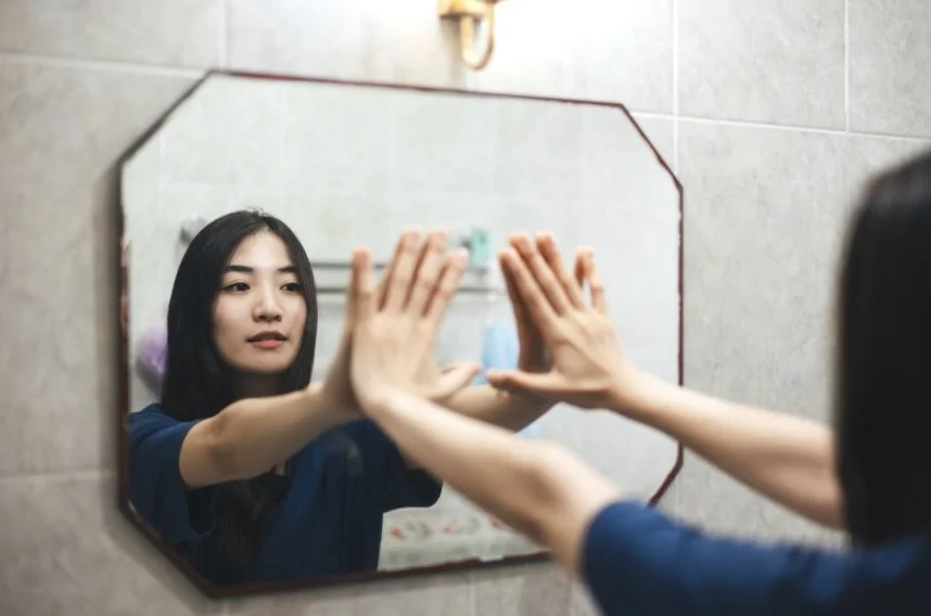 A young woman with long black hair looking at her reflection in a bathroom mirror, touching the mirror with both hands.