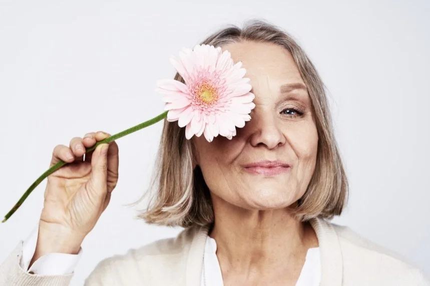 Older woman with shoulder-length gray hair holding a pink Gerbera daisy to her right eye, smiling softly.