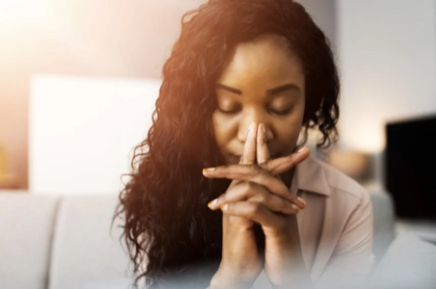 A woman with long curly hair sits with her eyes closed, hands clasped in prayer or contemplation