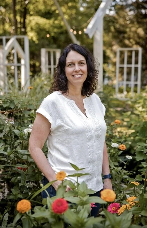 A woman with dark curly hair smiling while standing among flowering plants in a garden with a white trellis structure in the background.