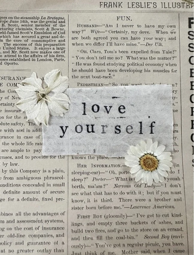 A newspaper page with a paper flower and a note that reads 'love yourself' placed on it.