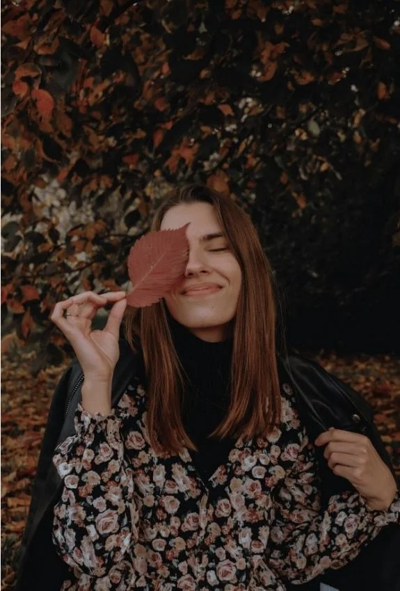 Young woman with long brown hair holding a large red autumn leaf in front of her eye, smiling with eyes closed, surrounded by fall foliage.