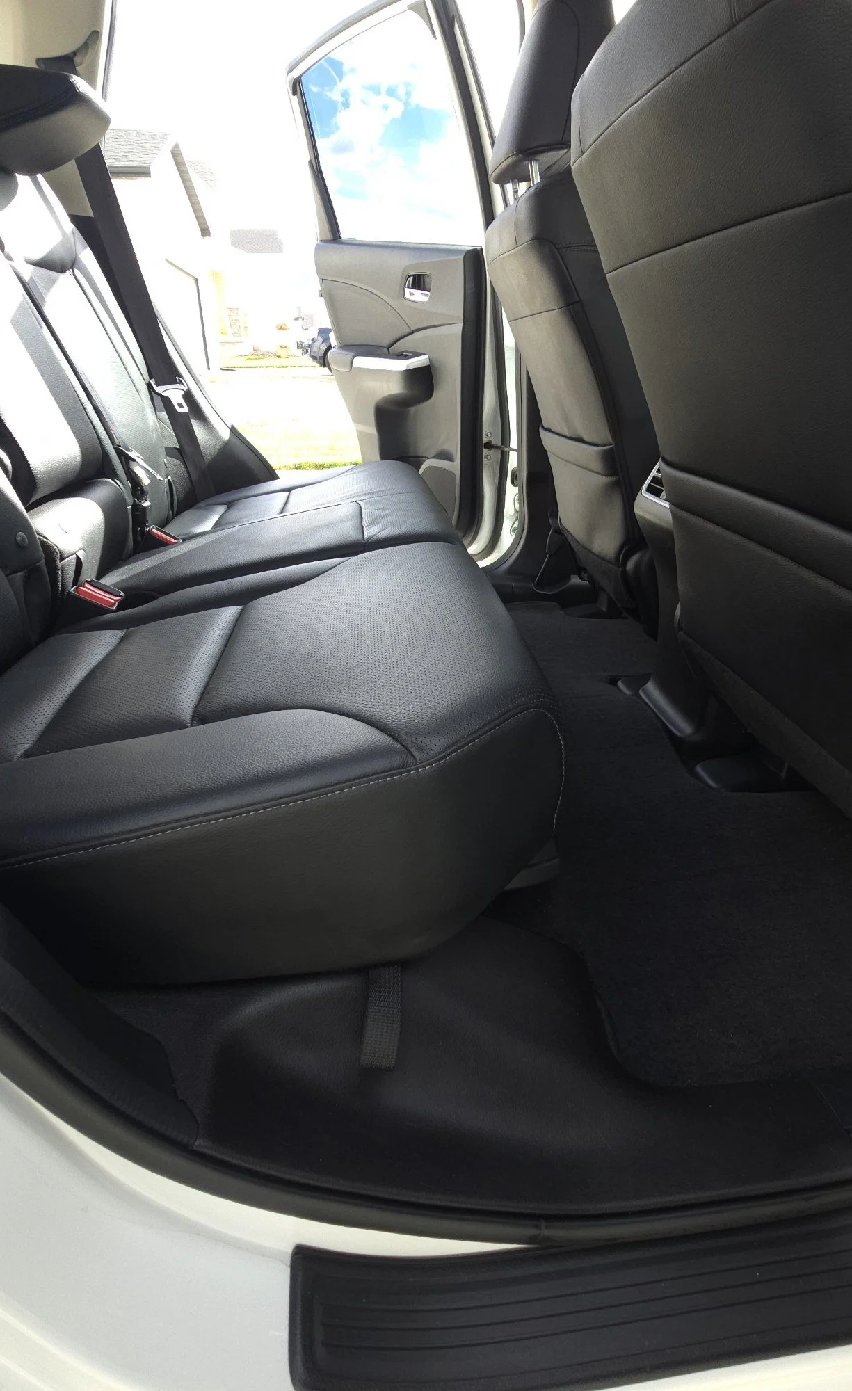The interior of a vehicle showing the back seat, with black leather upholstery, and open rear door with view outside of a parking lot and blue sky with clouds.