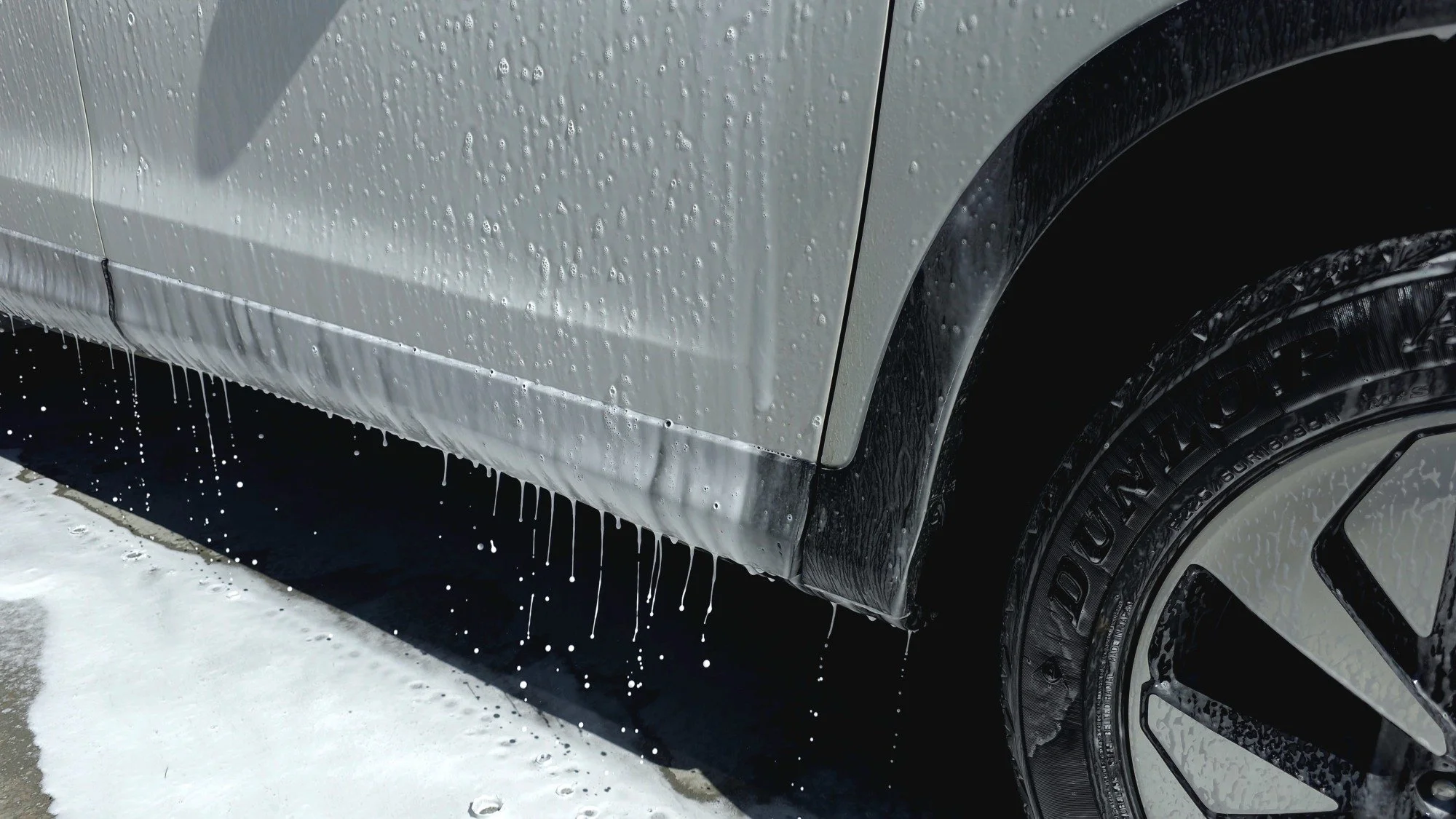 Close-up of a light-colored car's side, with water and soap suds dripping from the lower edge, washing in a car wash.