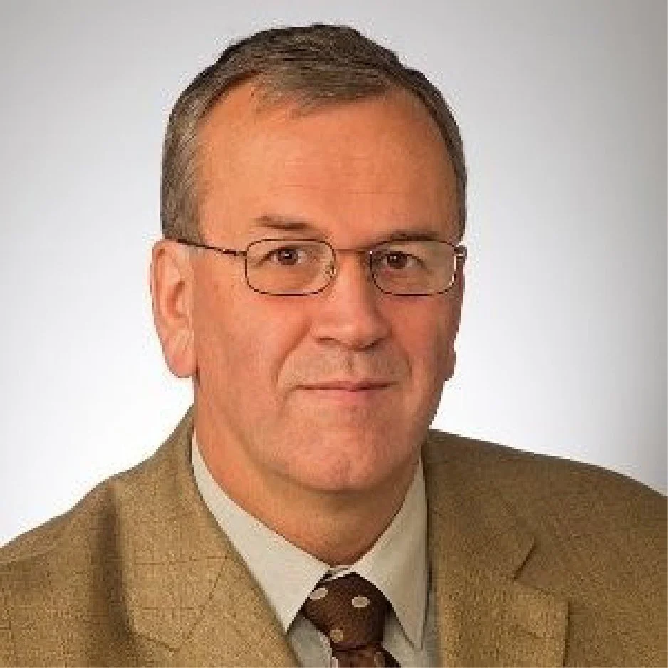 A professional middle-aged man with short brown hair and glasses, wearing a tan suit jacket, a white shirt, and a brown polka-dot tie, posing against a plain light gray background.