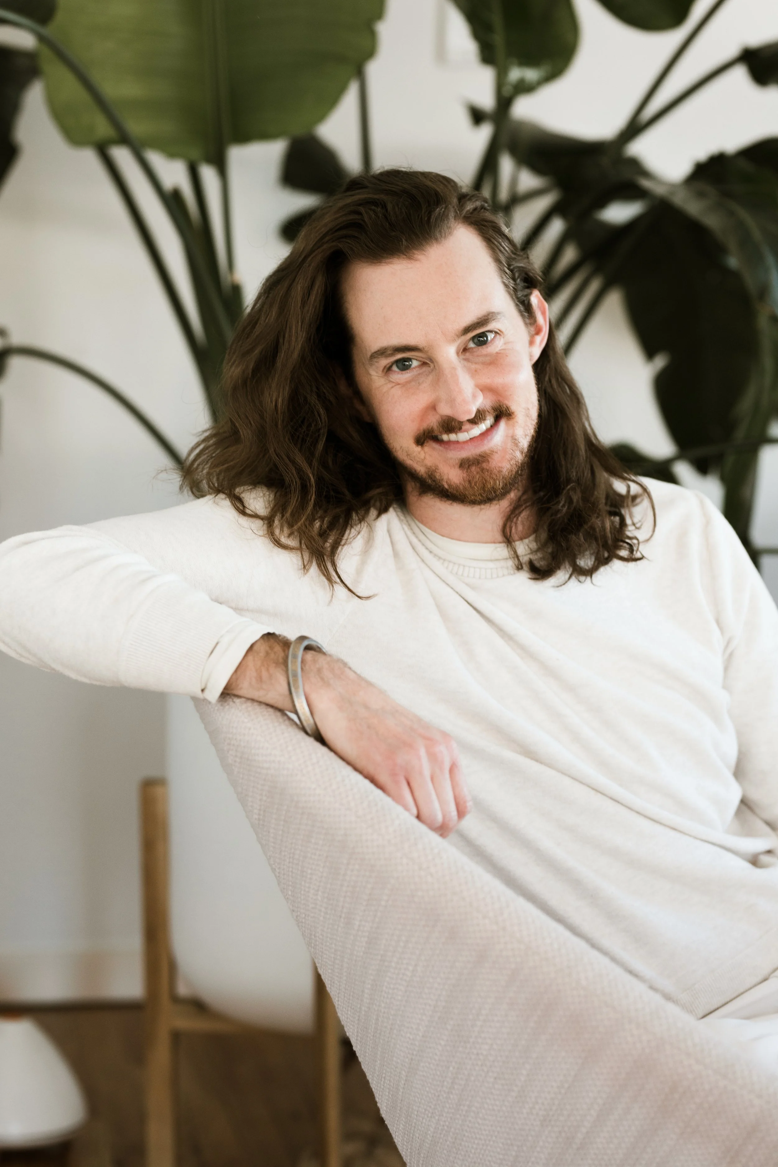 A man with long brown hair and a beard sitting in front of a large green plant, smiling at the camera, wearing a light-colored long-sleeve shirt.