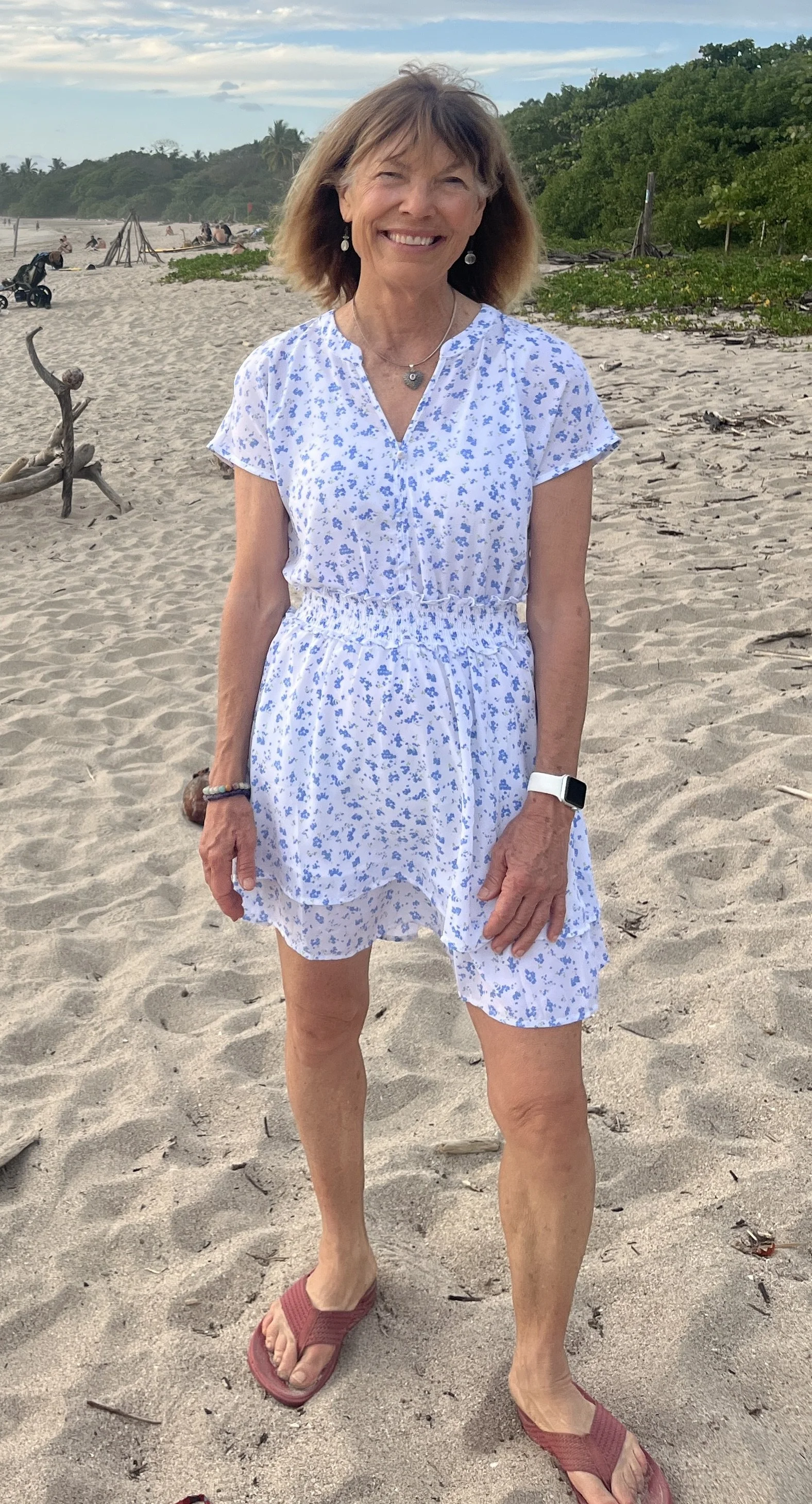 A smiling woman standing on a sandy beach with green foliage and blue sky in the background. She is wearing a white dress with blue floral patterns, red slide sandals, an Apple Watch, earrings, and a necklace.