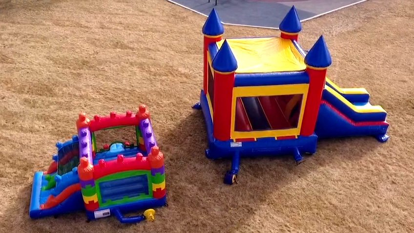 Aerial view of two inflatable rentals side by side on grass: a smaller, colorful toddler combo and a larger castle-style bounce house with a slide.