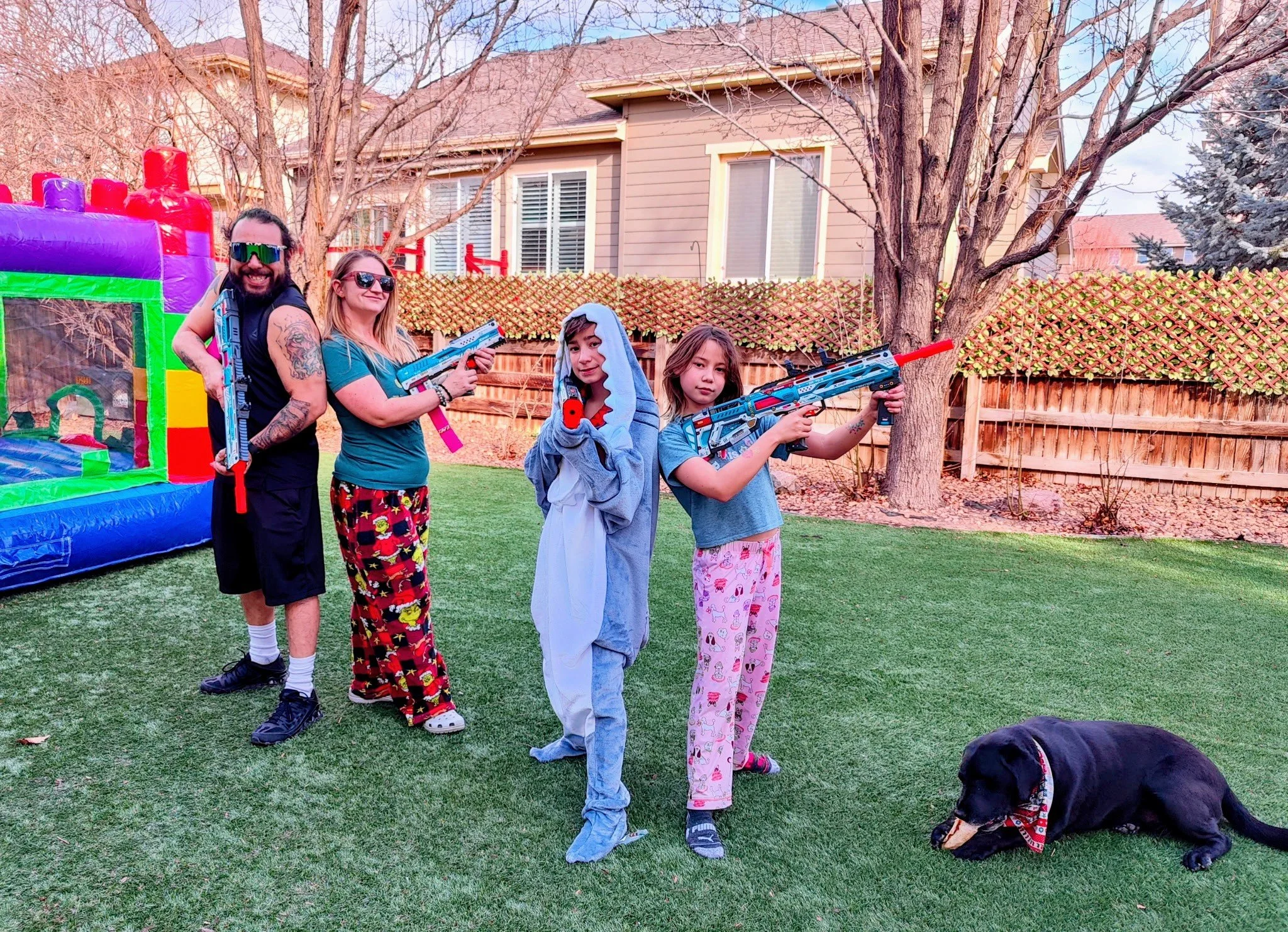Family of five with two children in pajamas, adults holding toy guns, a dog lying on the grass, and an inflatable bounce house in a backyard with trees and house in the background.