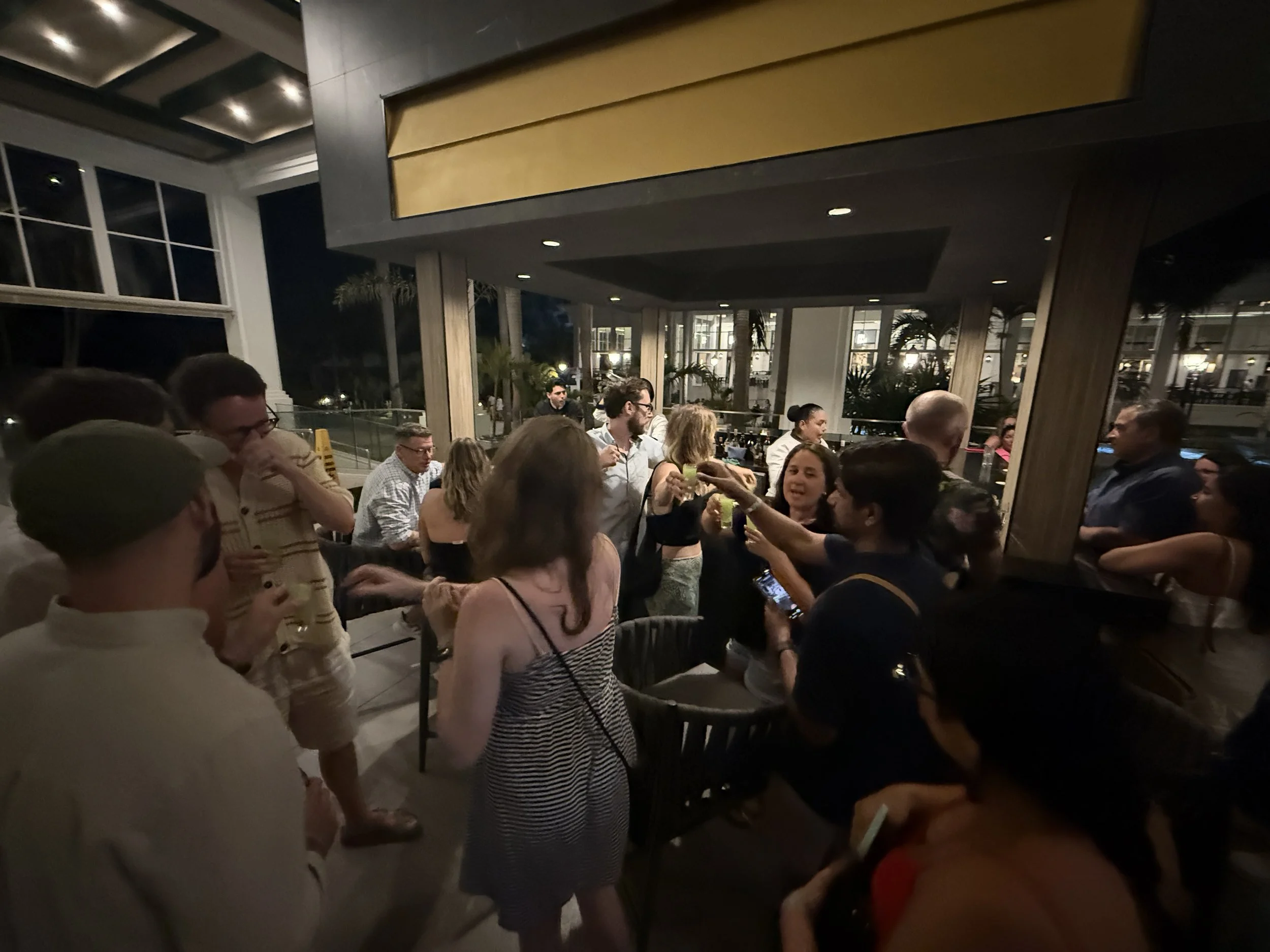 Group of people socializing at an indoor evening gathering, some holding drinks, in a modern room with large windows and outdoor view.