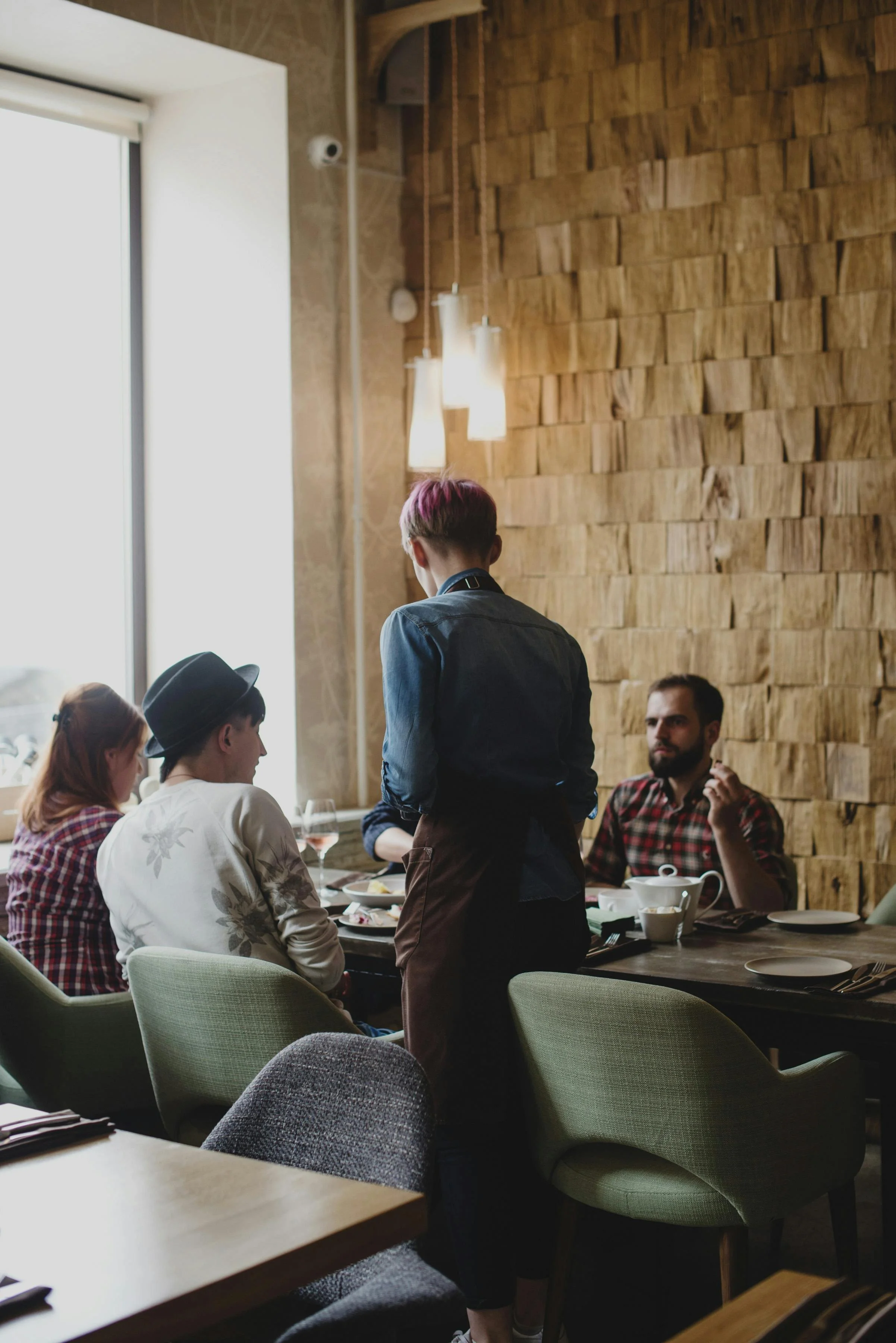 Group of people dining in a restaurant, server taking order, wooden wall decor, natural light from window.
