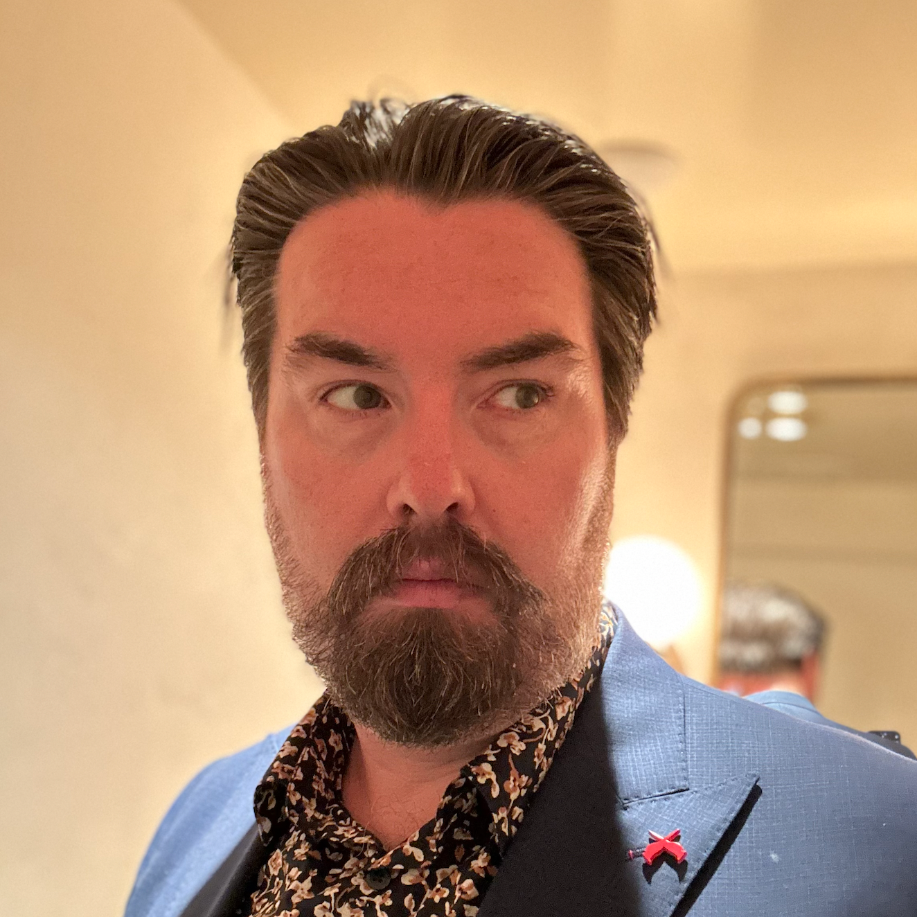 A man with dark hair and a beard, wearing a blue blazer and a patterned shirt, looking to the side in a warmly lit room.