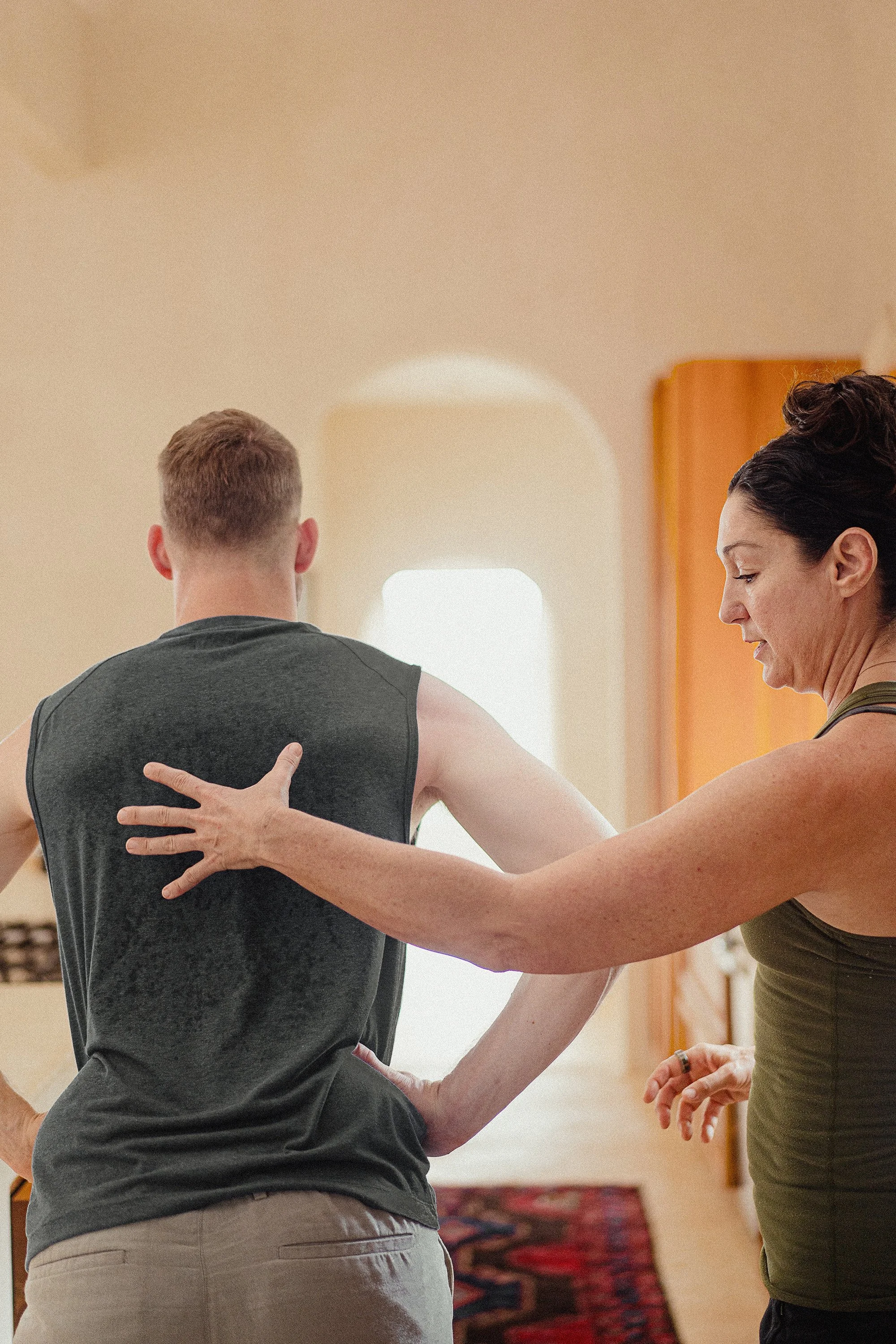 A woman is giving physical therapy to a man, helping him stretch his shoulder inside a room with natural light and warm-colored walls.