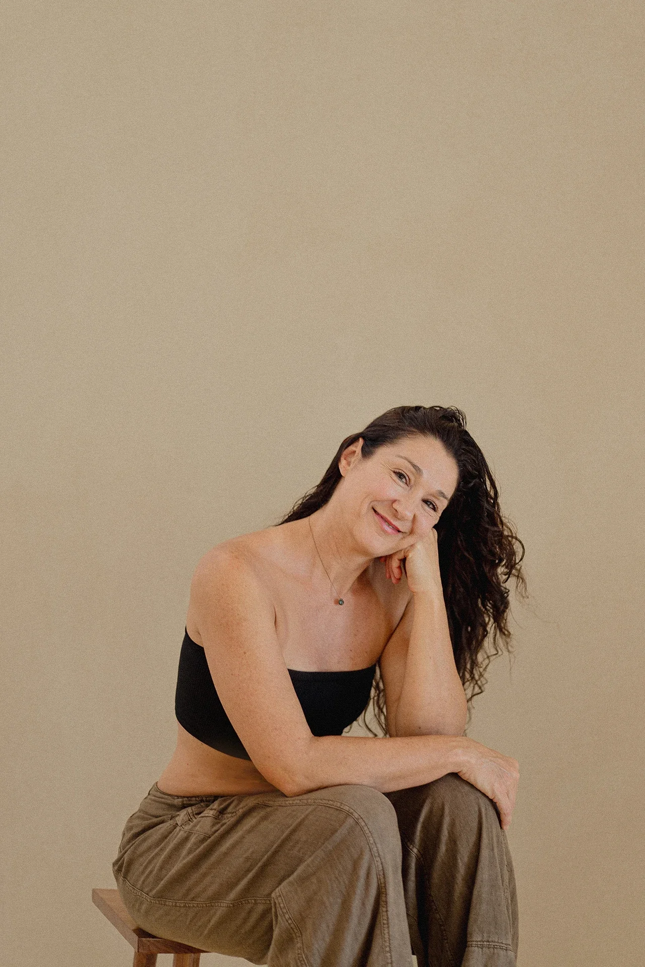 A woman with long, curly dark hair sitting on a wooden stool against a plain beige background, smiling with her head resting on her hand, wearing a black strapless top and brown pants.