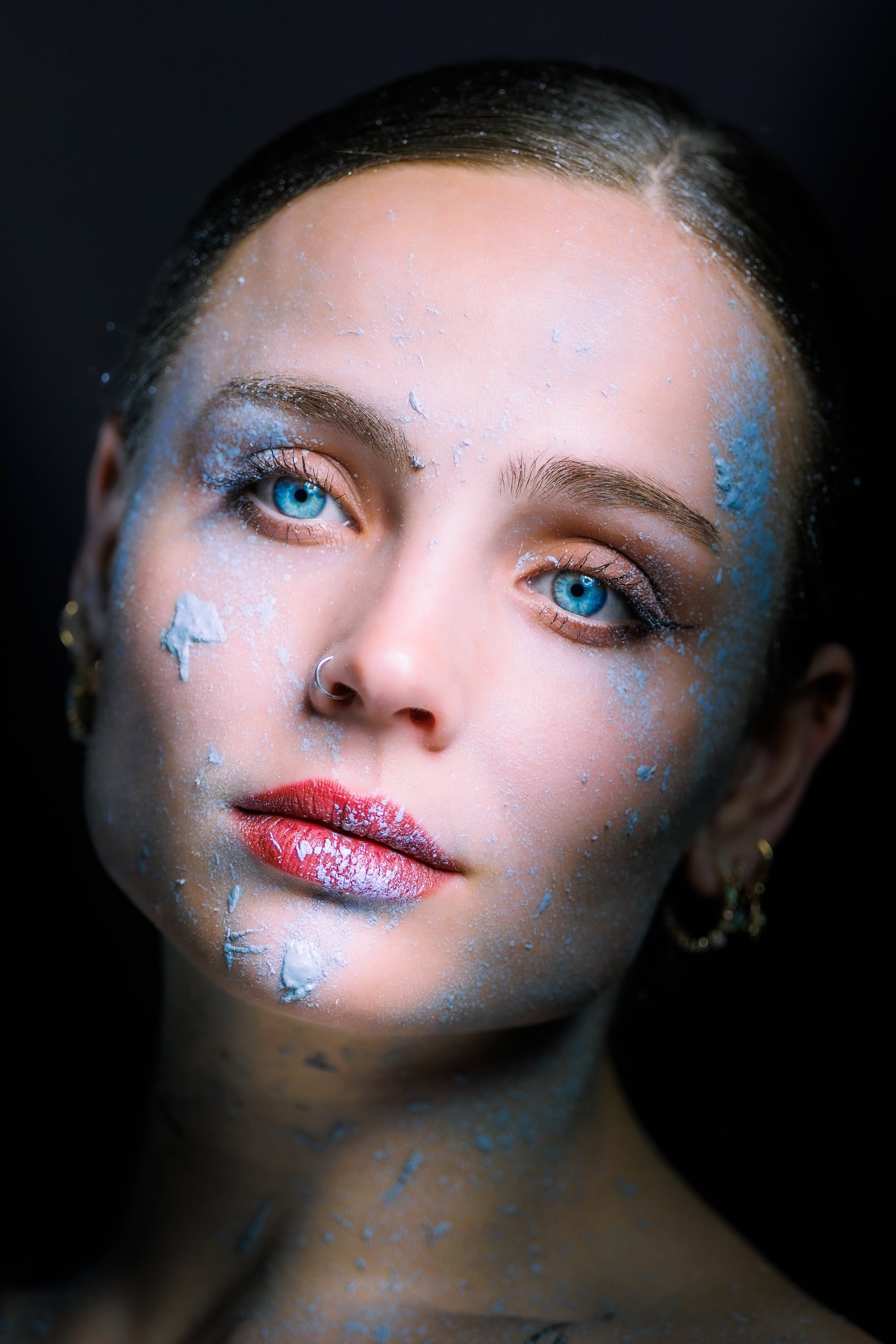 Nahaufnahme eines Gesichts einer Frau mit blauer Augen, nassen Haaren und blauer Farbe auf Haut und Lippen, schwarze Hintergrund.