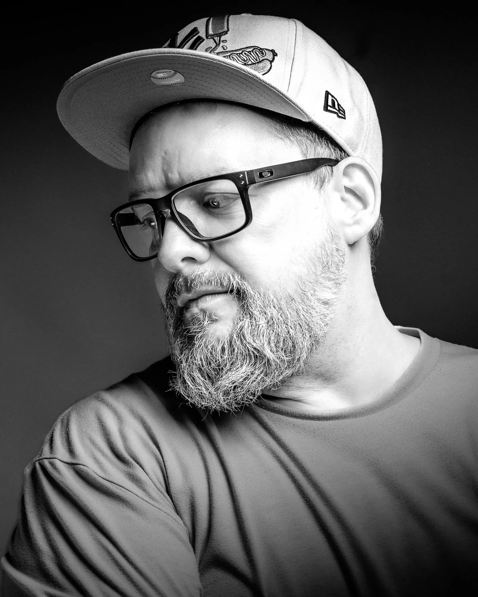 Schwarz-weiß-Foto eines Mannes Vollbart, trägt eine Baseballkappe und schwarze Brille, blickt seitlich nach unten.