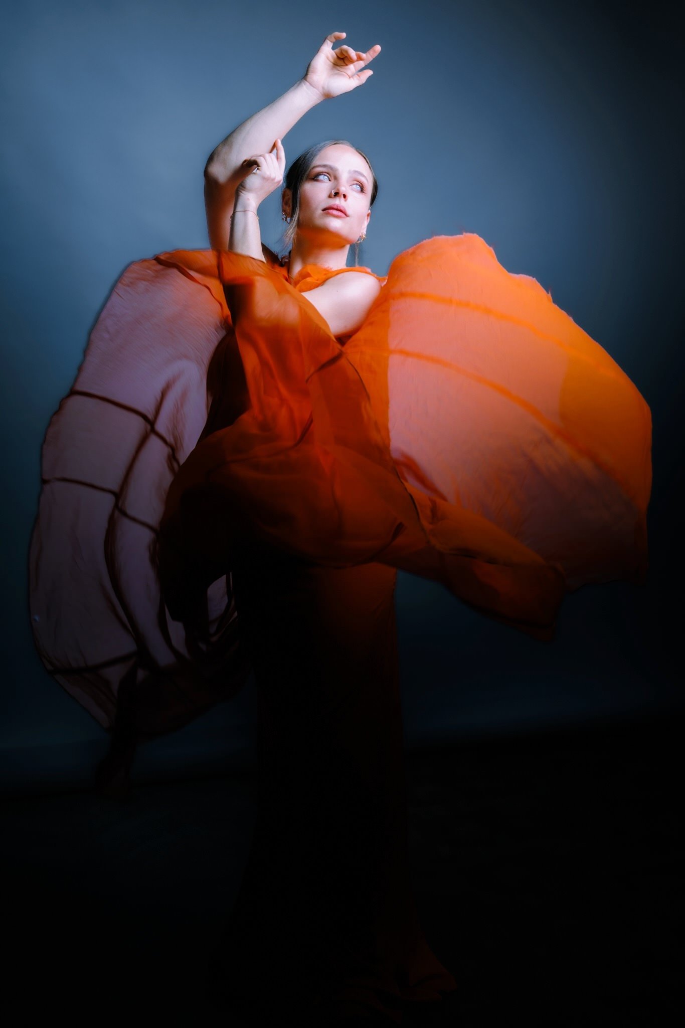 Frau in einem orangefarbenen Kleid mit Blick nach oben, vor einem dunklen Hintergrund fotografiert.