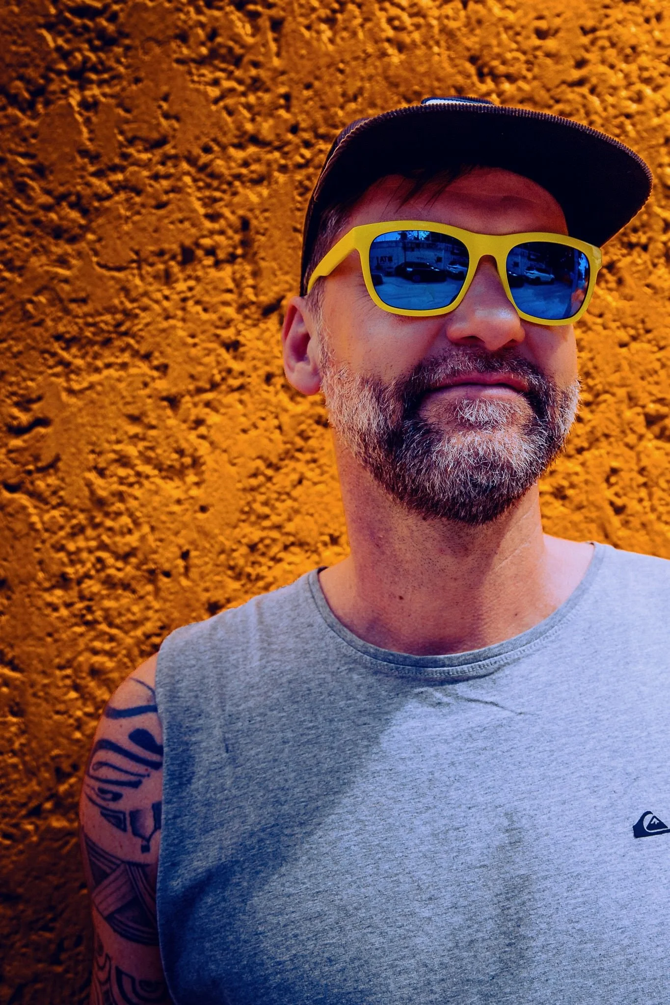 Porträt eines Mannes mit grauem Bart, schwarzen Haaren, schwarzen Baseballcap, gelben Sonnenbrillen mit blauer Reflexion, grauem Tanktop, Tattoo am linken Arm, vor einer orangfarbenen Wand.