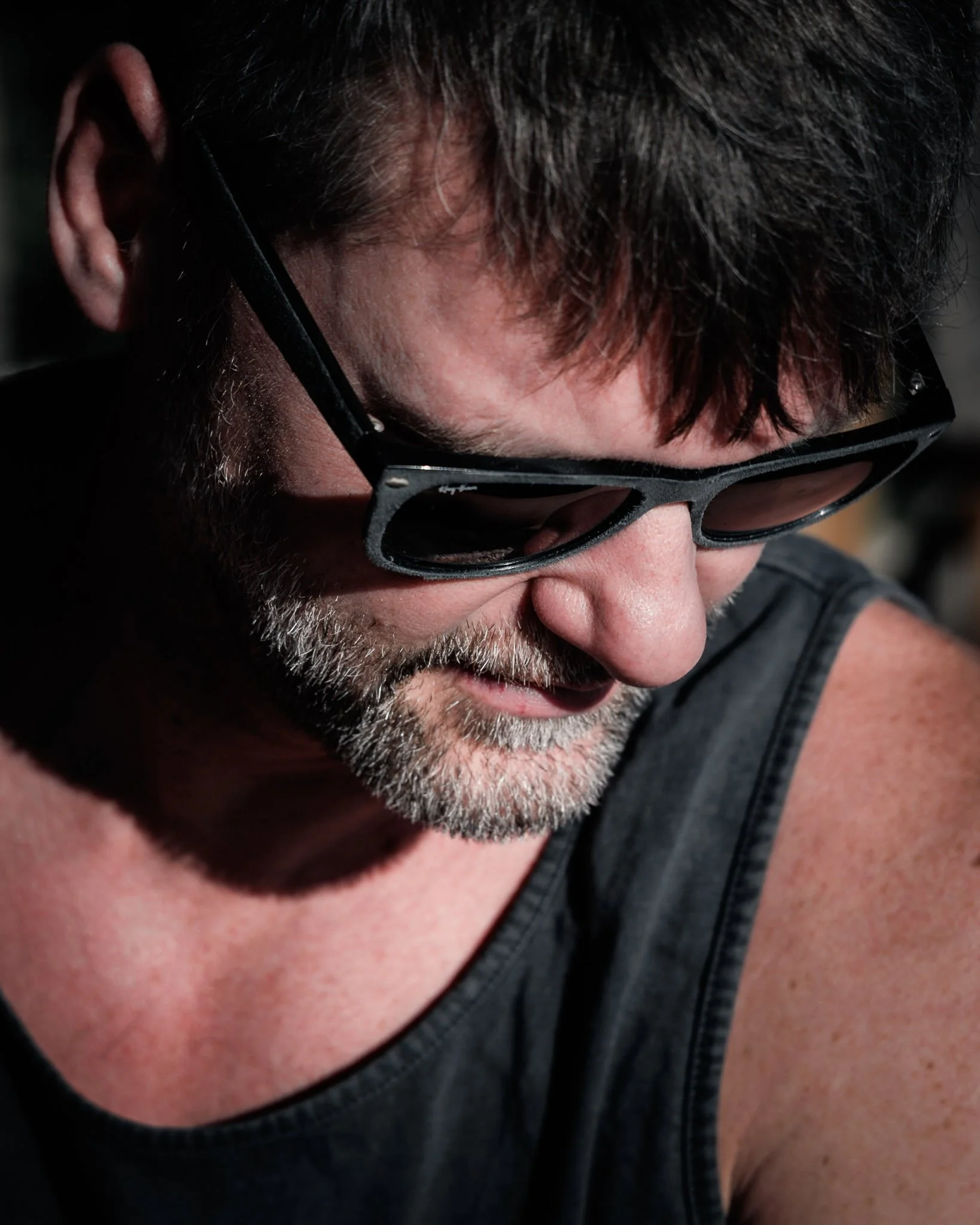 Nahaufnahme eines Mannes mit dunklem Haar und Bart, trägt schwarze Sonnenbrille, trägt ein dunkles T-Shirt, sichtlich im Schatten.