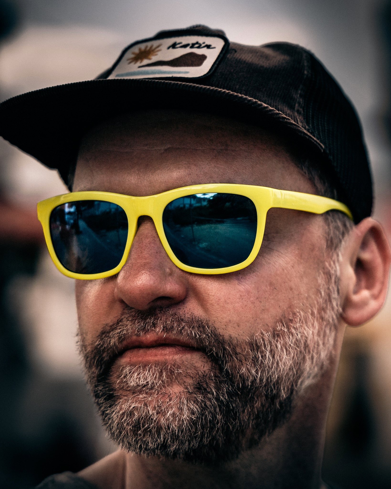 Nahaufnahme eines Mannes mit grauem Bart, der eine gelbe Sonnenbrille trägt und eine schwarze Kappe mit einem Patch auf dem Kopf trägt.