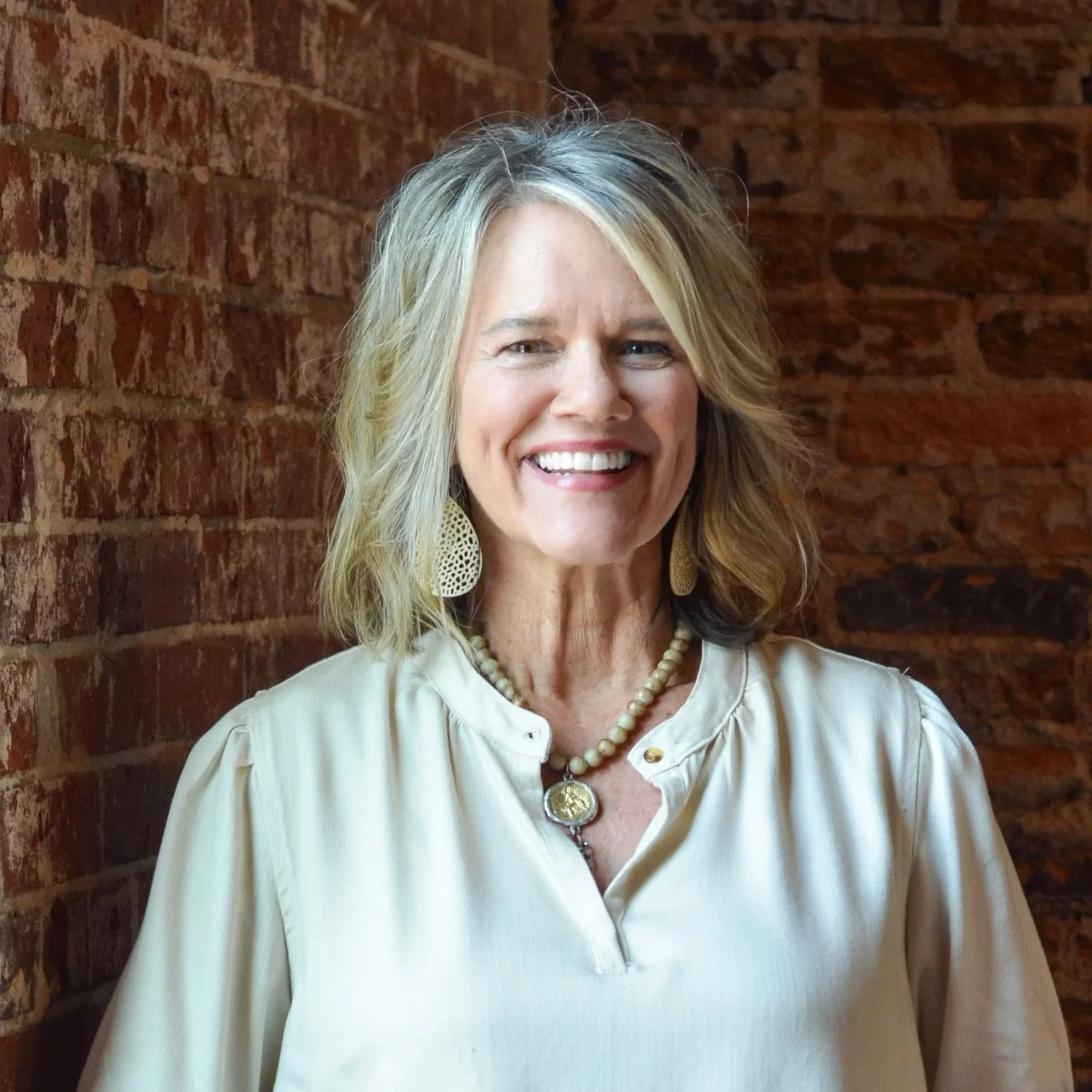 Smiling middle-aged woman with blonde hair wearing a white eyelet blouse and large earrings, standing outdoors in front of a house with greenery.