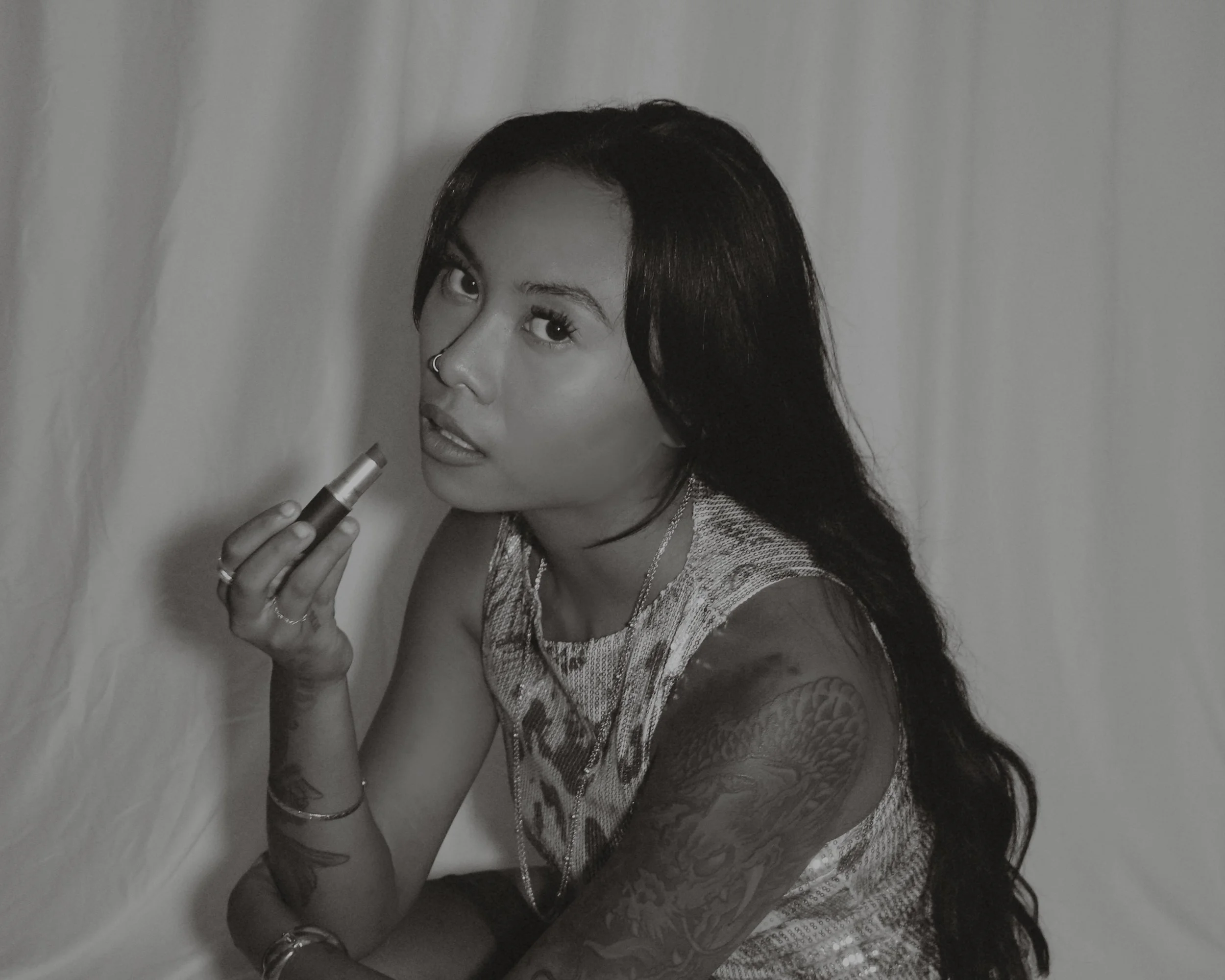 A woman with long dark hair and tattoos on her arms is holding a lipstick near her lips. She has a nose piercing and is wearing a sleeveless patterned top, jewelry, and is looking at the camera against a plain background.