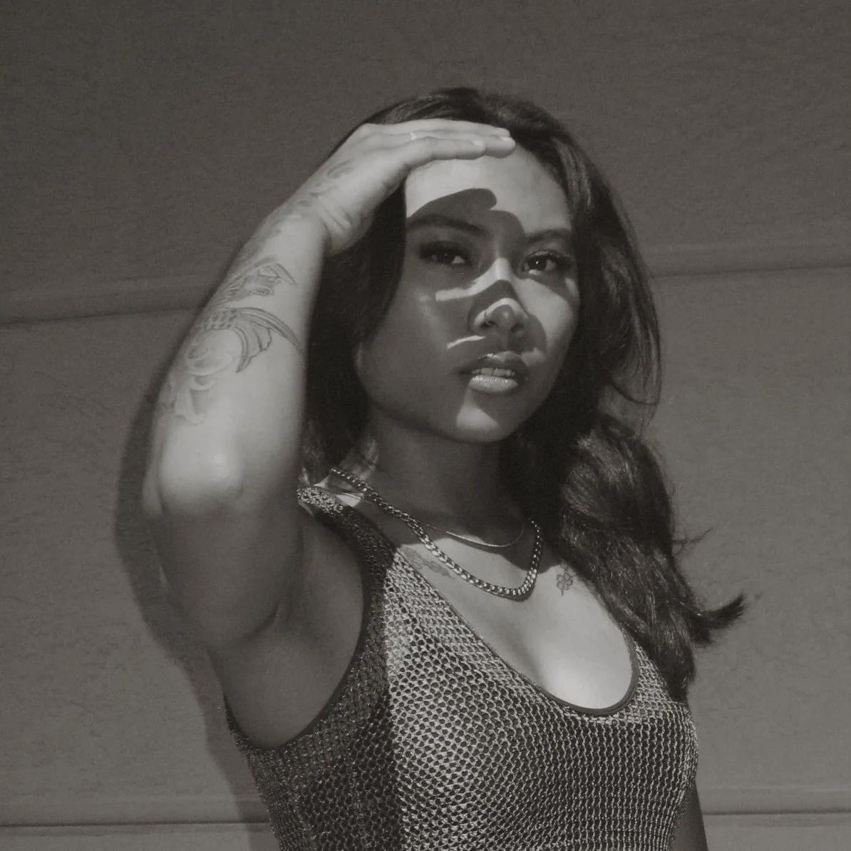 A woman with long wavy hair, tattoos on her arm, wearing a sleeveless top and layered necklaces, posing with her hand on her forehead, in black and white.