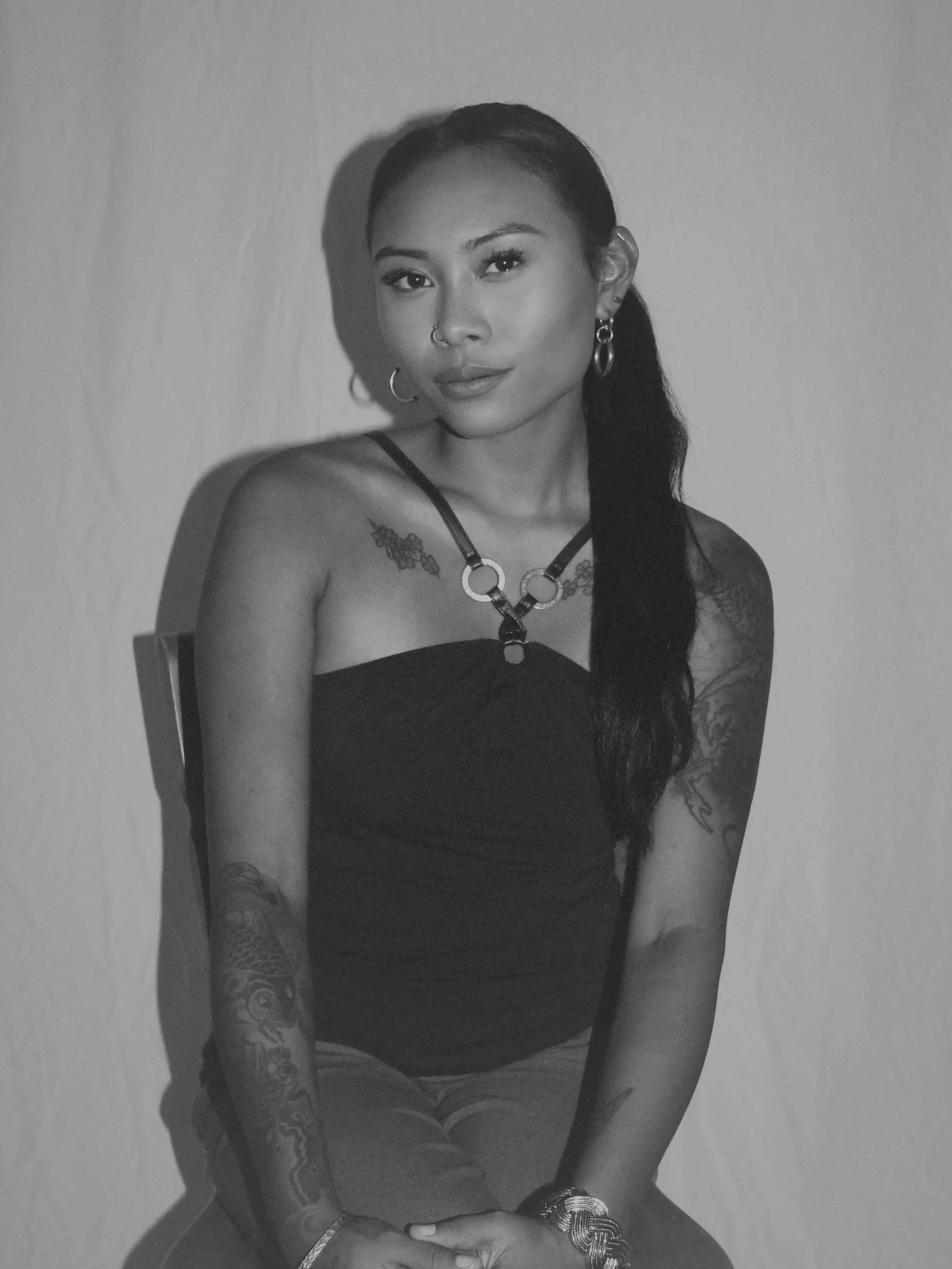 A young woman with tattoos, earrings, and a necklace, sitting against a plain wall, looking at the camera, in a black strapless top.