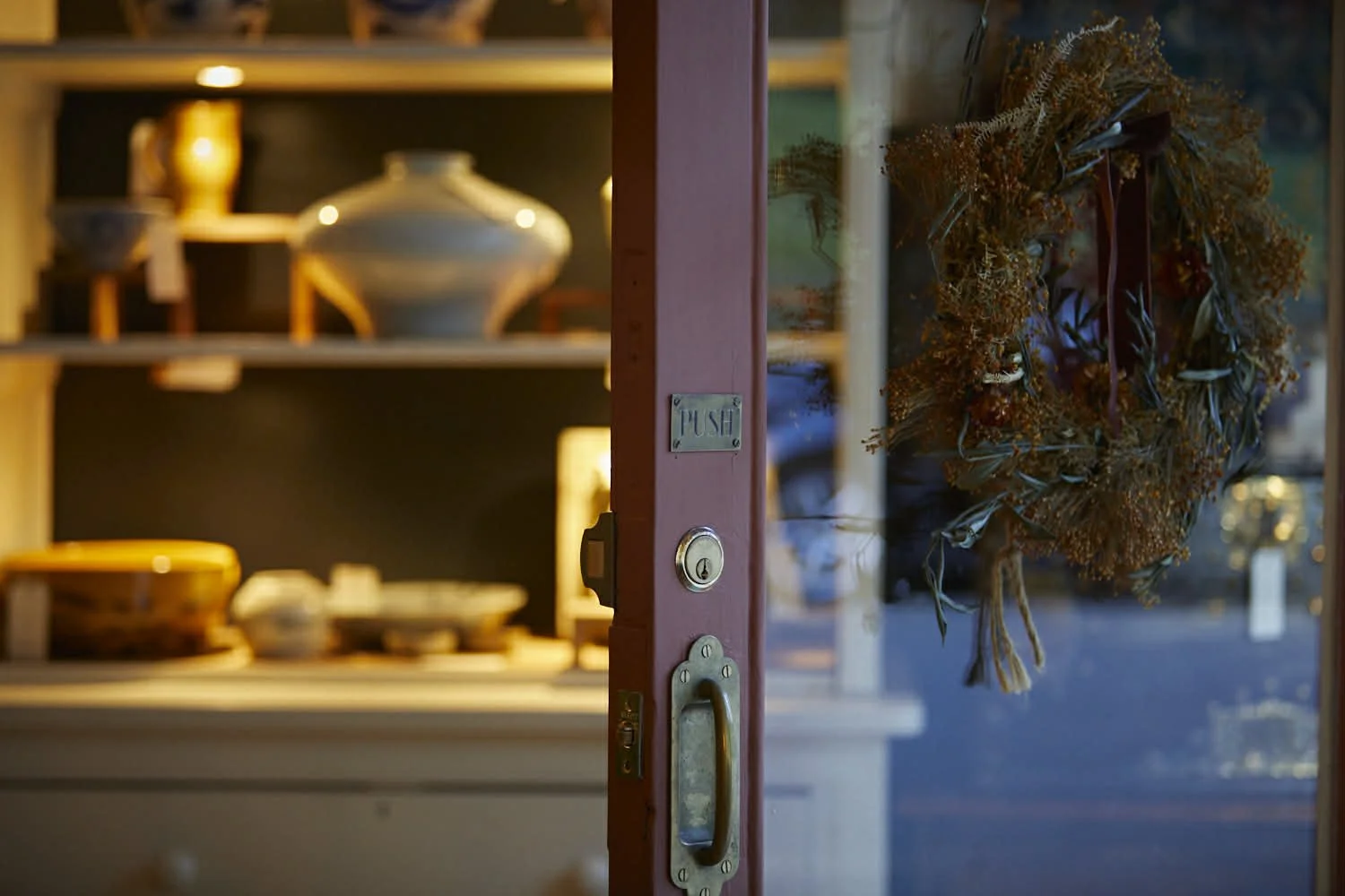 A glass door with a dried floral wreath hanging on it and a brass handle, revealing a cozy shop interior with shelves of pottery and ceramics.