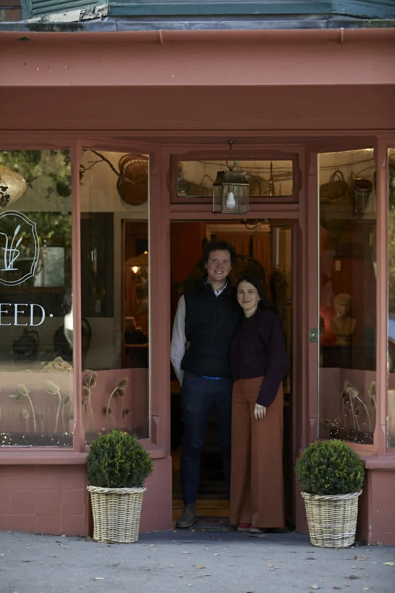 Two women standing in the doorway of a pink storefront, smiling for the camera. Potted plants are outside, with decorative items visible inside the shop.