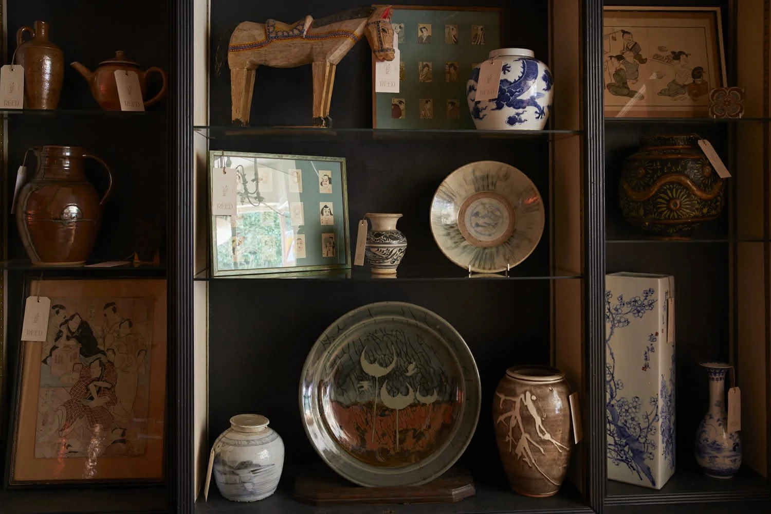 Display of various decorative Asian pottery and art pieces arranged on black shelves, including vases, bowls, framed artwork, and carved wooden figurines.
