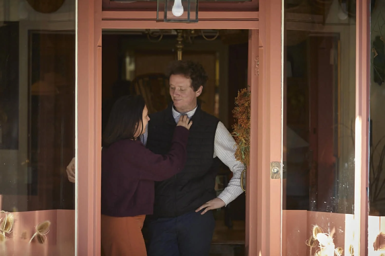 A woman is adjusting a man's bow tie through a doorframe, with a cozy indoor setting visible behind them.
